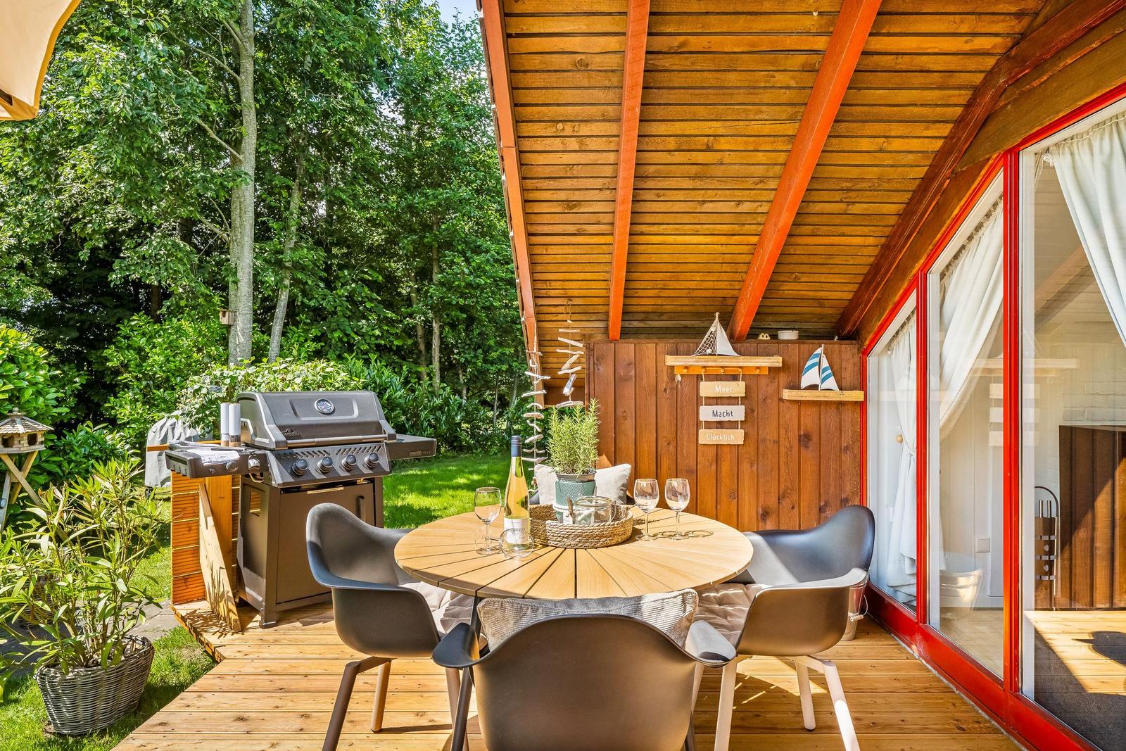 Terrasse mit Grill, Tisch und Stühlen. Holzverkleidung und Fenster mit roten Rahmen.