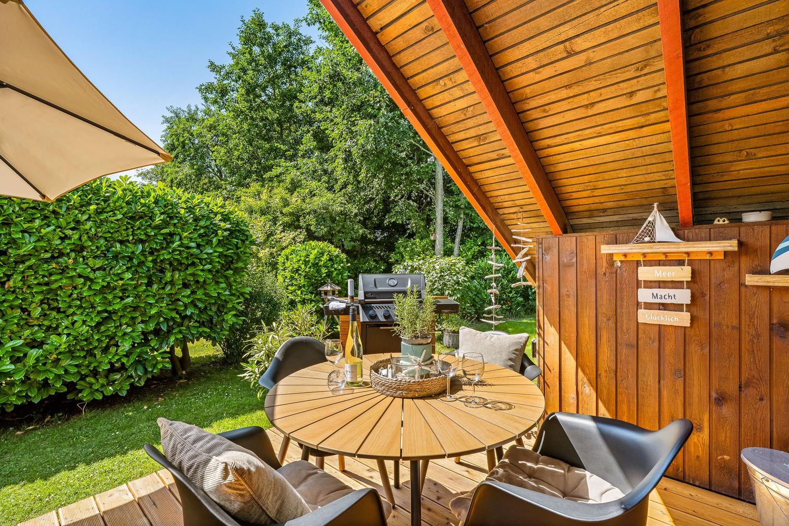 Terrasse mit Holztisch, Stühlen, Grill und grüner Umgebung unter Holzdecke.