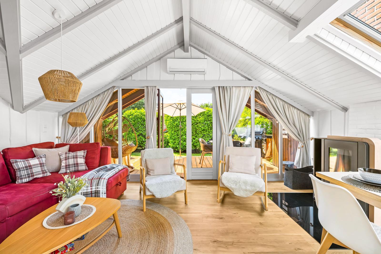 Ein Wohnraum mit rotem Sofa, zwei Sesseln und großem Fenster zum Garten.