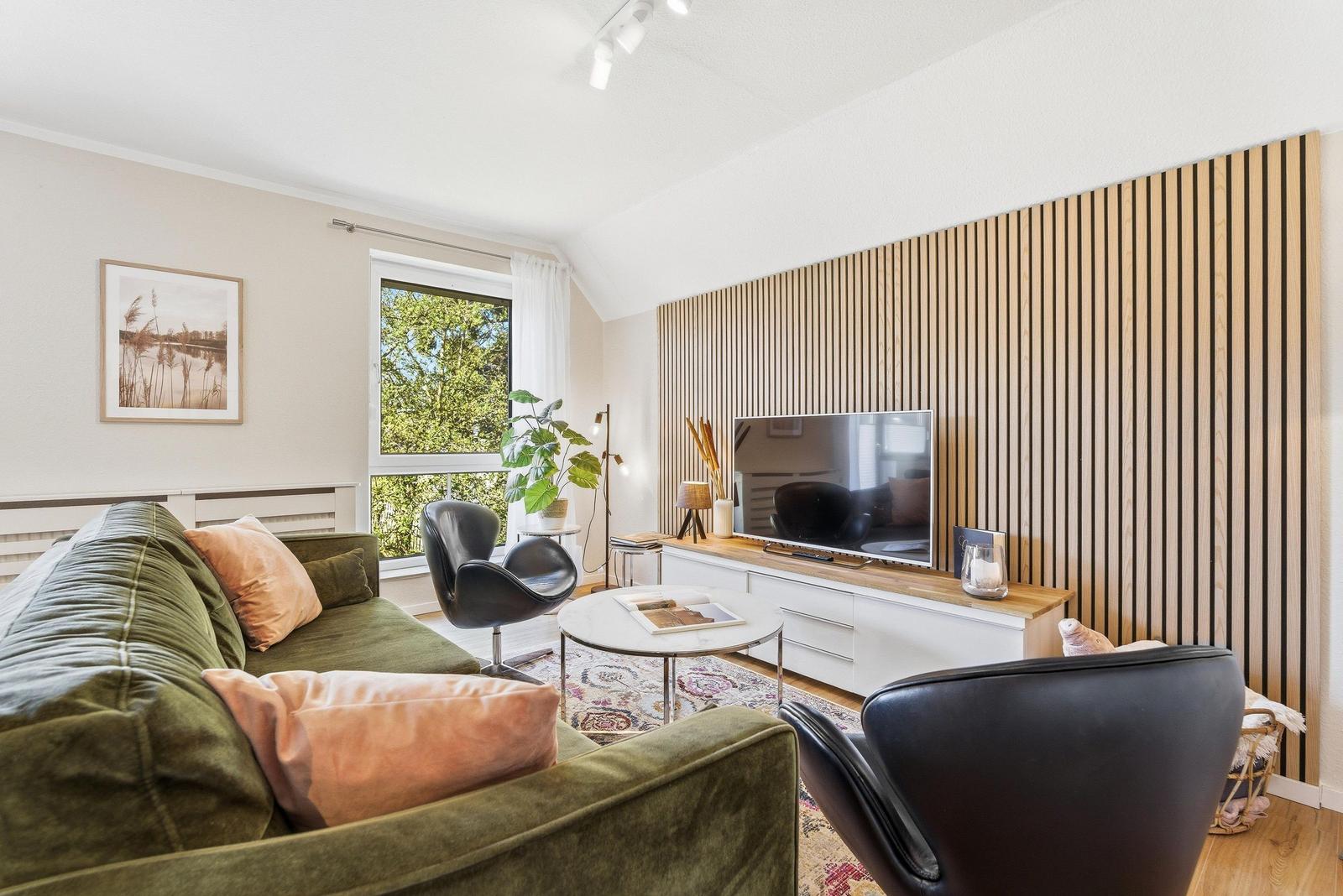 Wohnzimmer mit grünem Sofa, schwarzem Sessel und Holzwand. Fenster mit Blick auf Bäume.