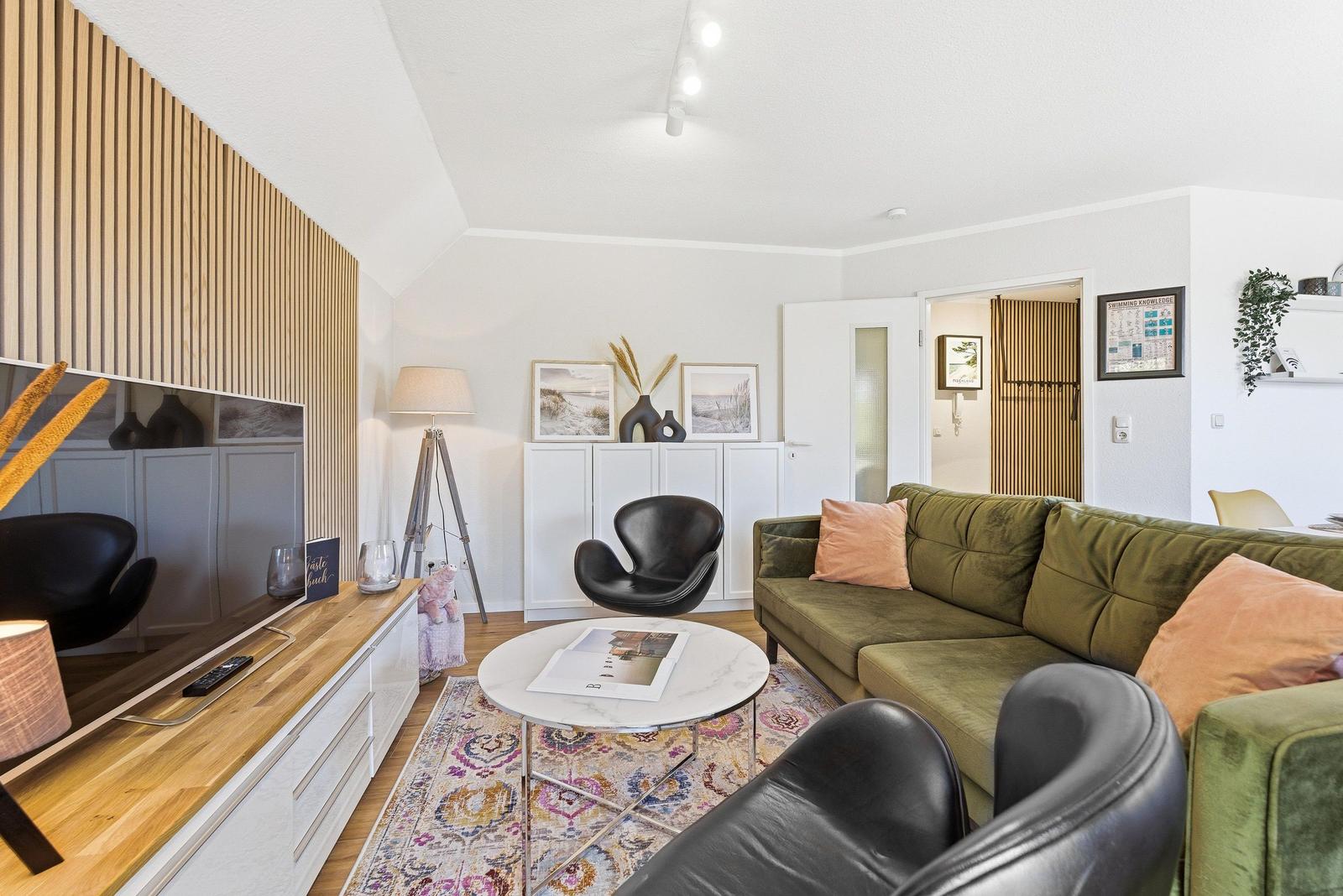 Wohnzimmer mit grünem Sofa, schwarzen Sesseln und Holzwand. Runder Tisch auf Teppich.