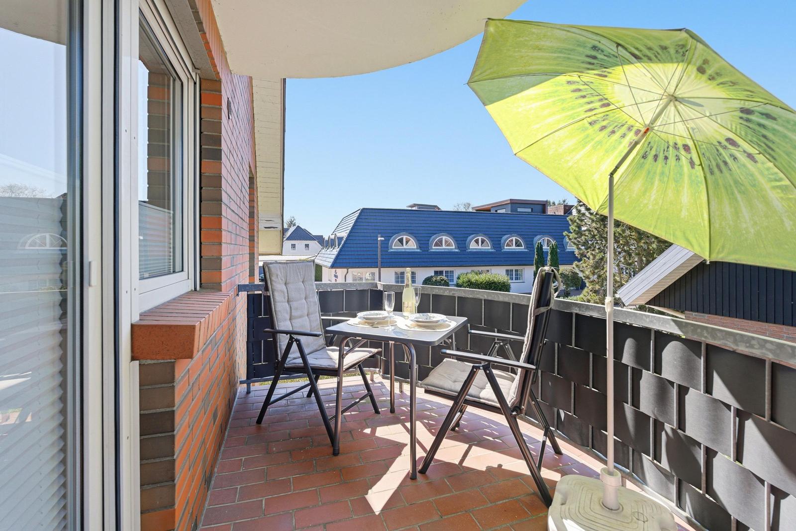 Terrasse mit Tisch, zwei Stühlen und grünem Sonnenschirm. Blick auf Haus mit blauem Dach.