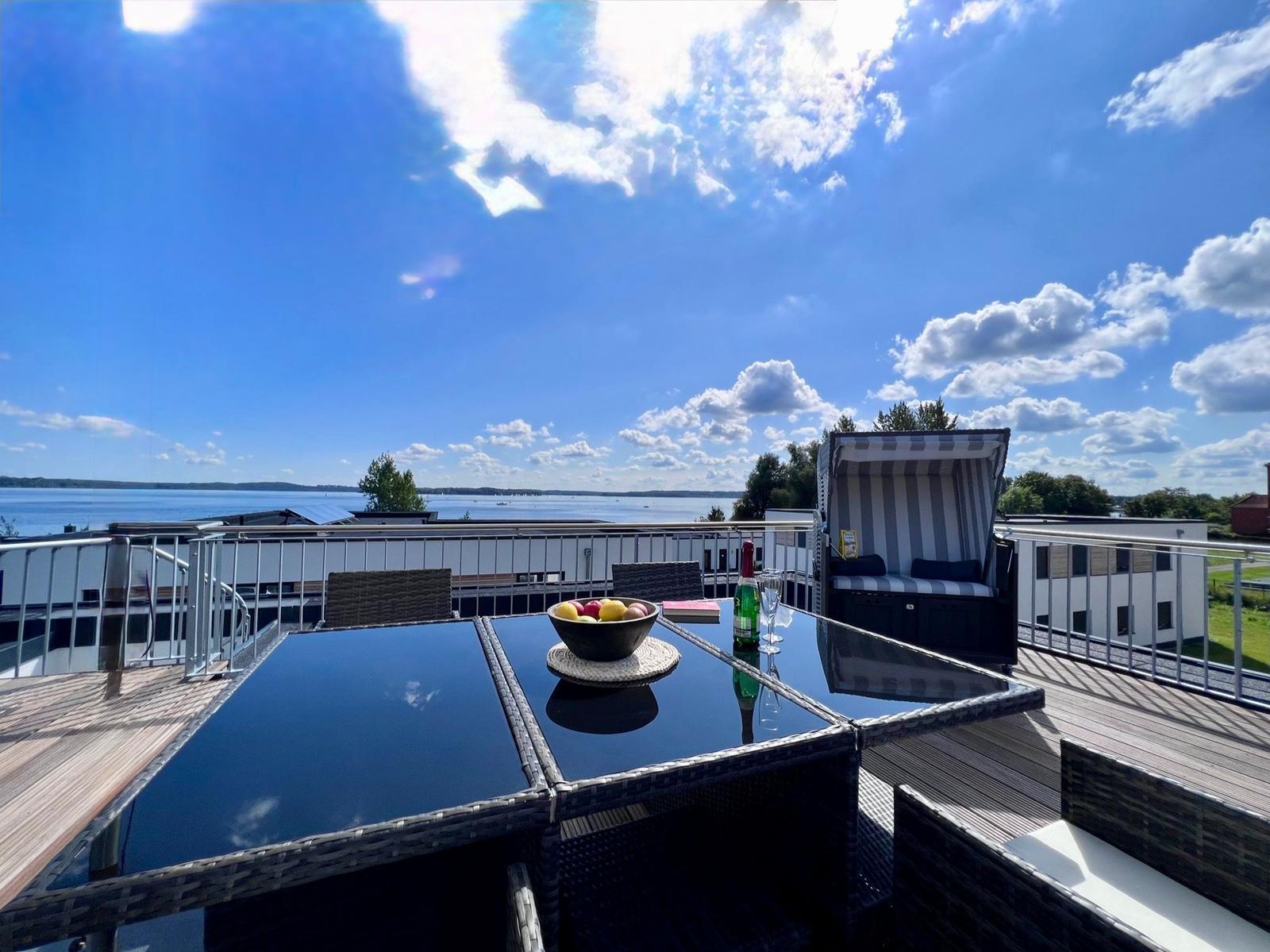 Terrace with table, chairs, and lounge under blue sky with water view.