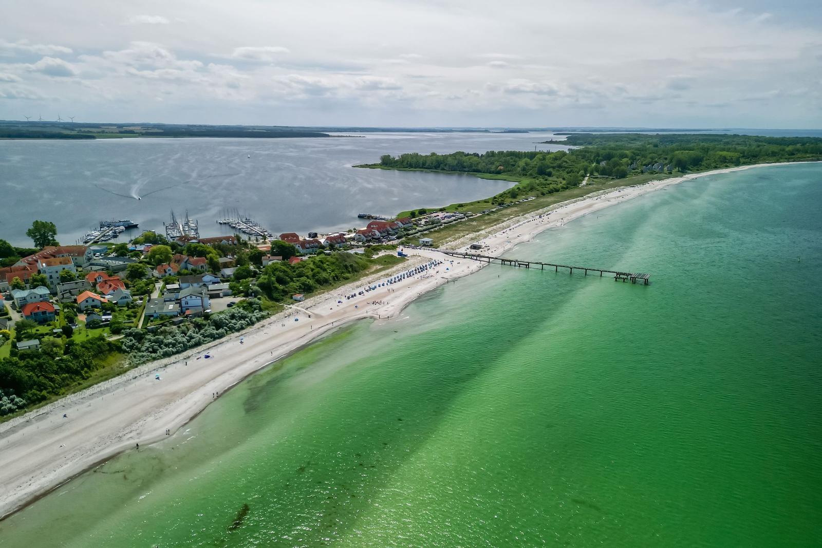 Aerial view of a beach with turquoise water, wooden pier, and coastal village with red-roofed houses.
