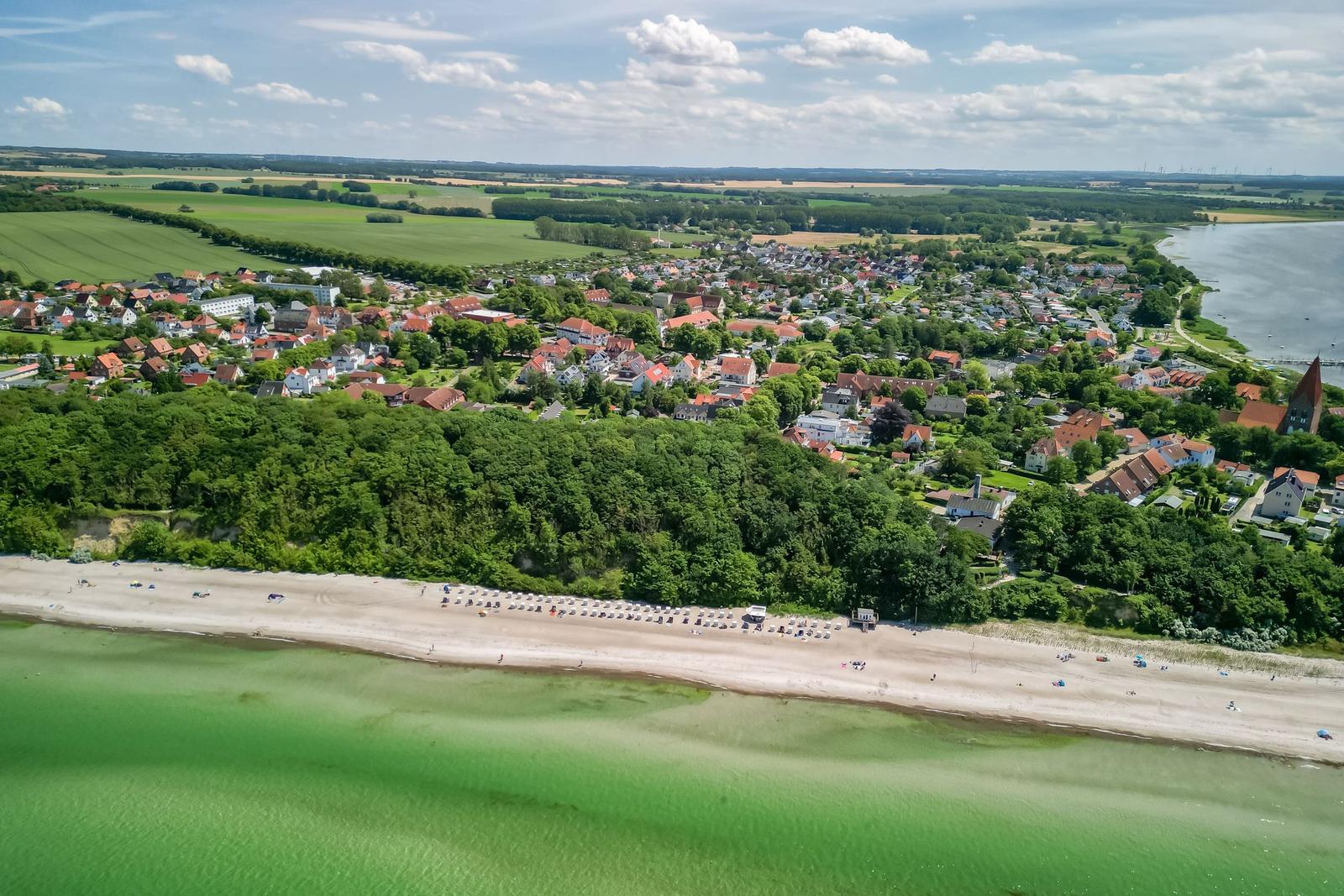 Strand mit Badegästen, grünes Wasser, Dorf mit Häusern und Wald im Hintergrund.