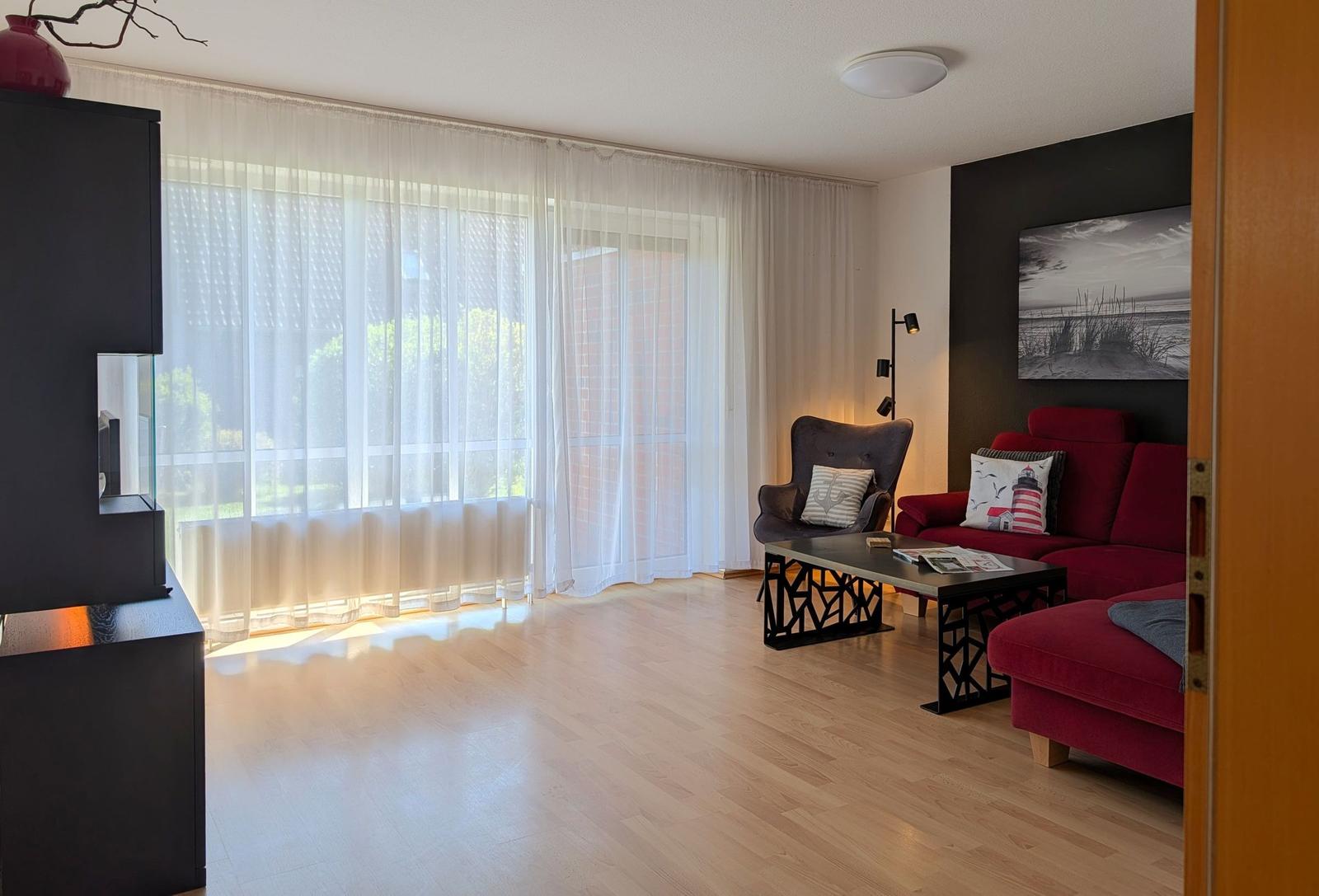 Wohnzimmer mit rotem Sofa, Sessel und Tisch. Große Fenster mit weißen Vorhängen.