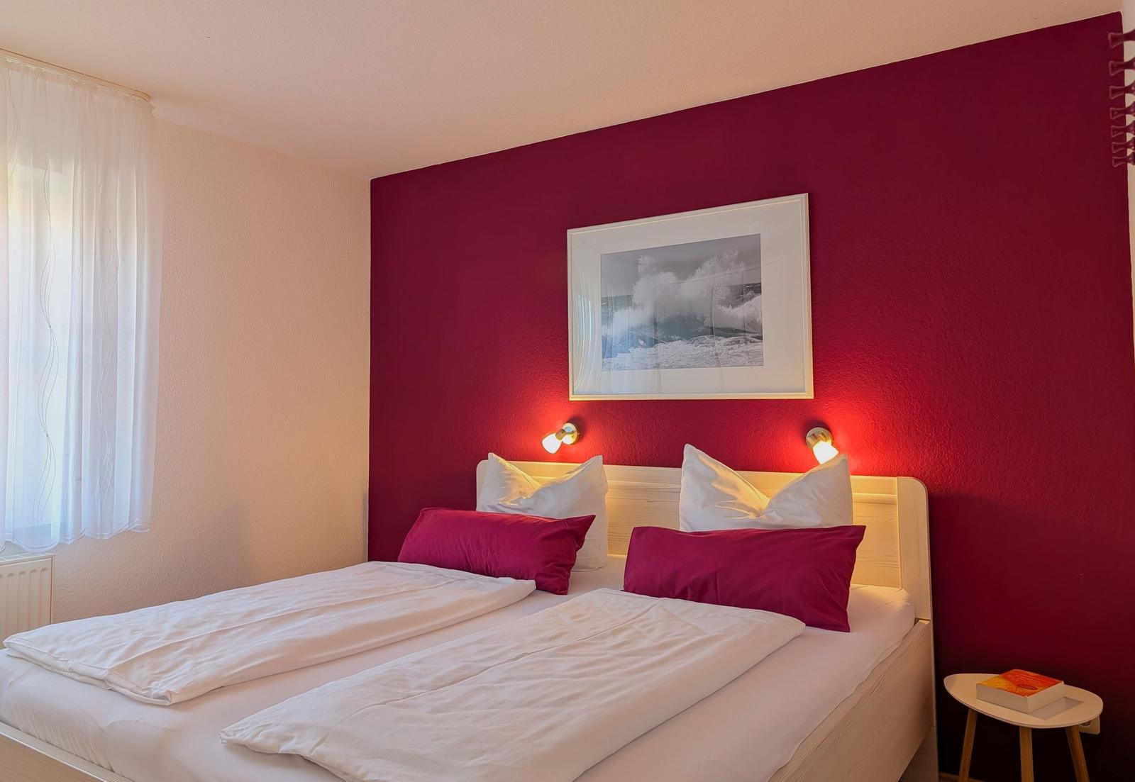 Ein Schlafzimmer mit roter Wand, Bett mit weißen und roten Kissen, Wandbild und Nachttisch.