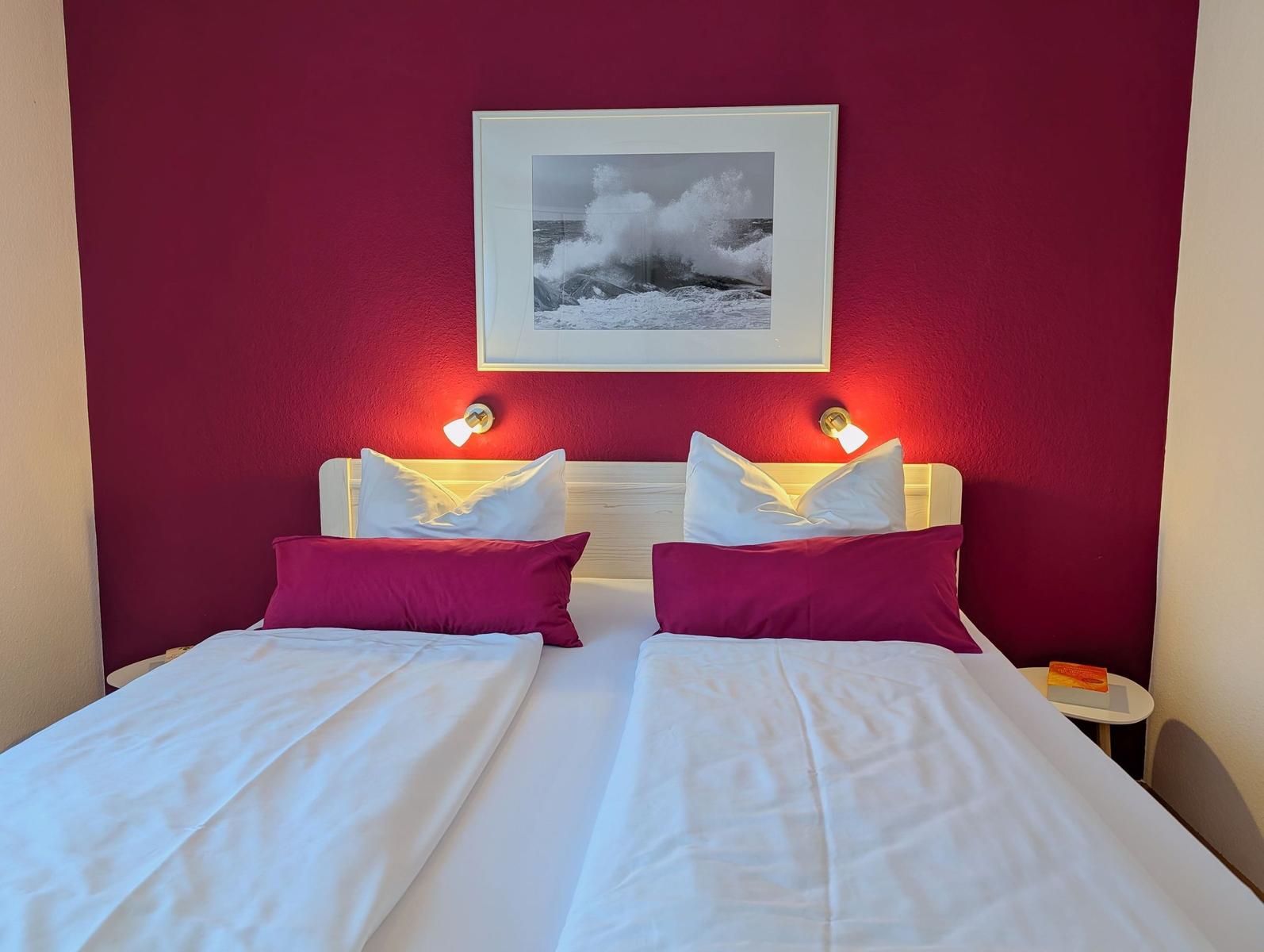 Zwei Betten mit weißen Laken und magentafarbenen Kissen an einer roten Wand mit Bild und Wandlampen.