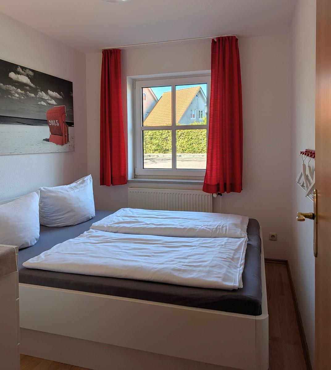 Schlafzimmer mit Bett, Fenster und roten Vorhängen. Wandbild und Heizkörper sichtbar.