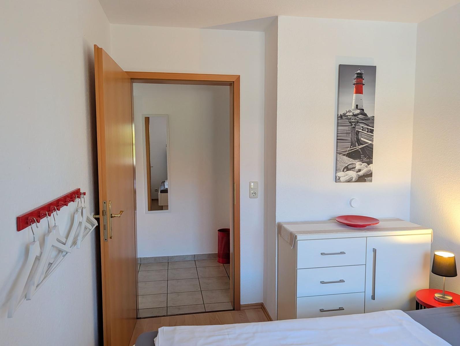 Schlafzimmer mit Kleiderhaken, Schrank und Bild an der Wand. Tür führt in einen Flur.