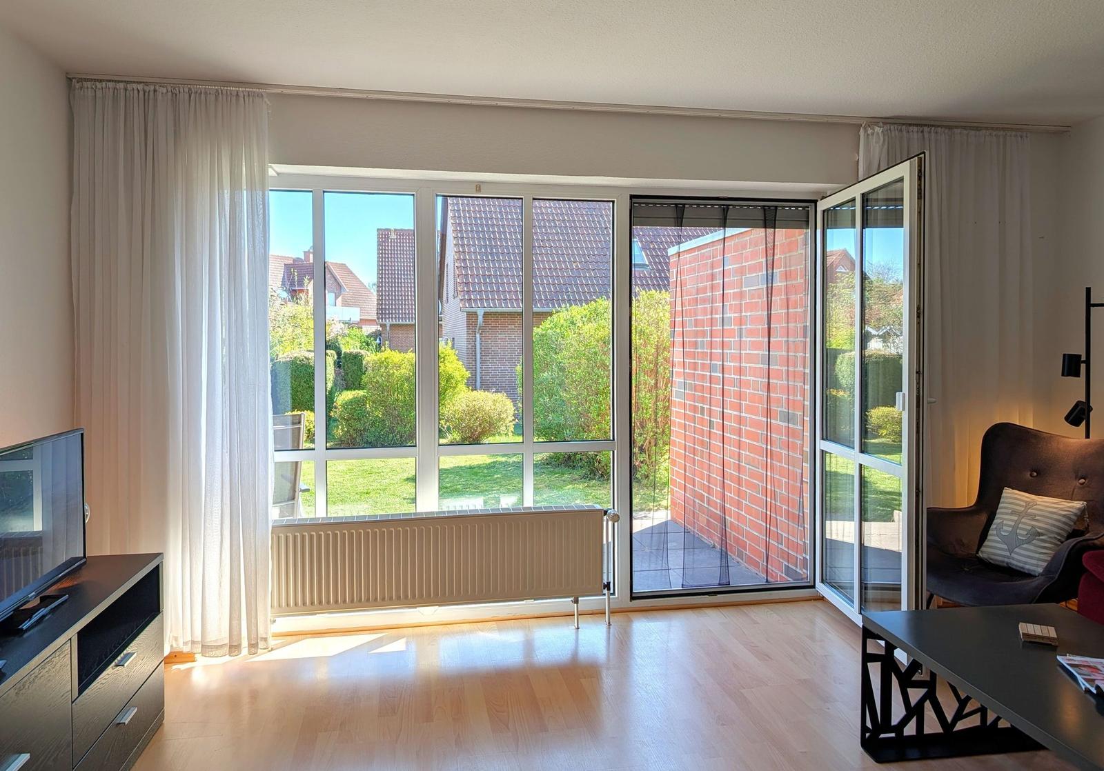 Wohnzimmer mit großem Fenster und Terrassentür, Blick auf Garten und Nachbarhäuser.