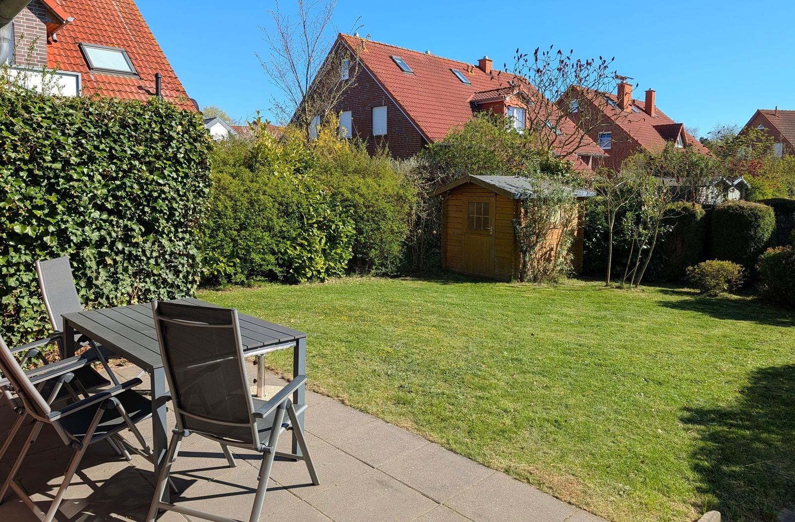 Garten mit Terrasse, Tisch, Stühlen und Holzschuppen. Hintergrund: Häuser mit roten Dächern.