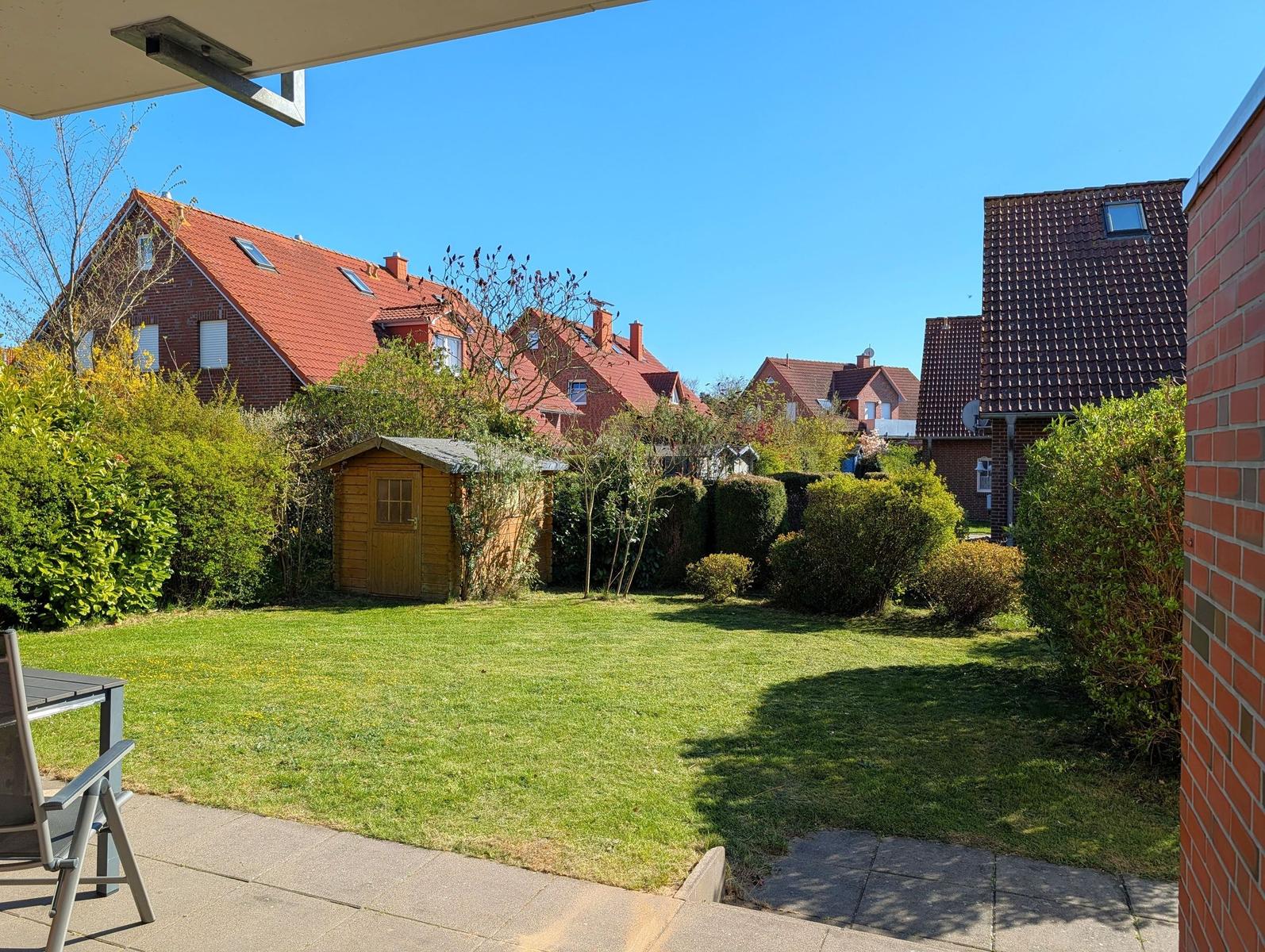 Garten mit Rasen, Holzschuppen und Sträuchern. Blick auf benachbarte Häuser mit roten Dächern.