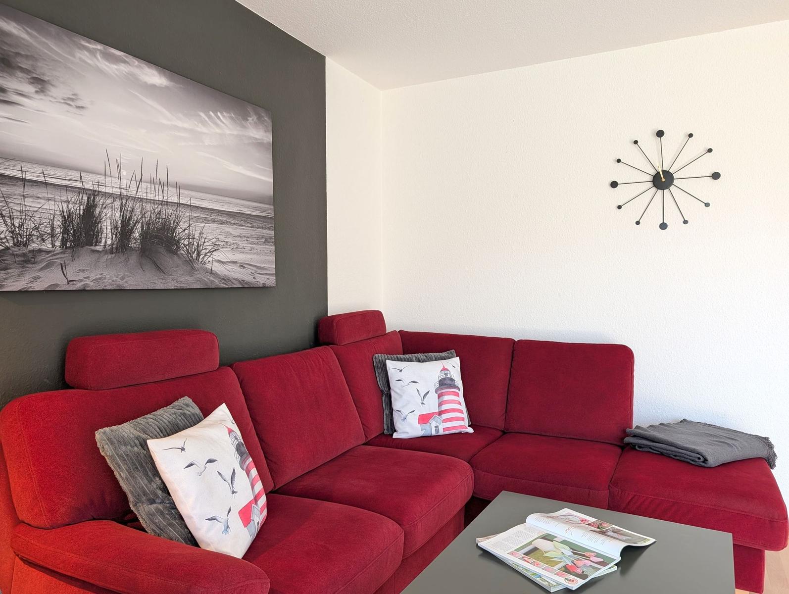 Roter Sofa mit Kissen, Wanduhr und Strandbild an der Wand.
