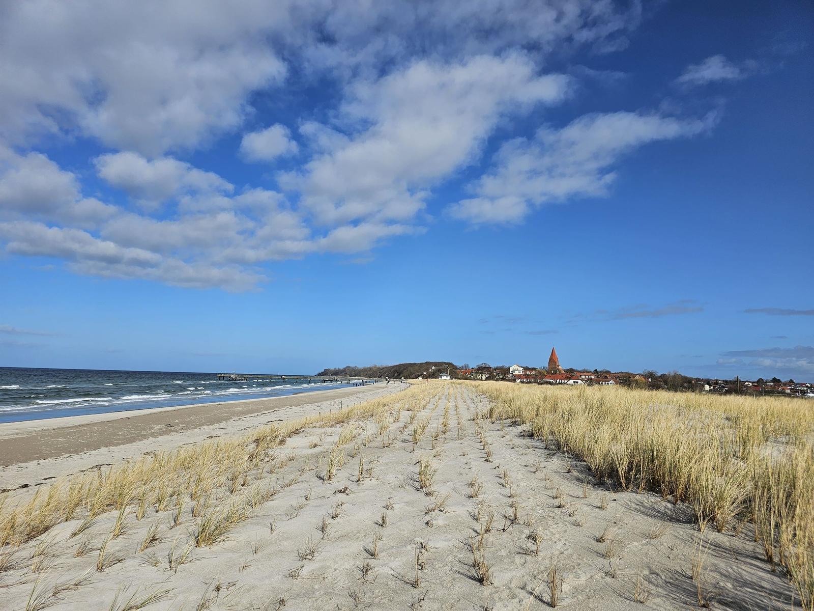 Strand mit Dünen, See und Dorf im Hintergrund unter blauem Himmel mit weißen Wolken.