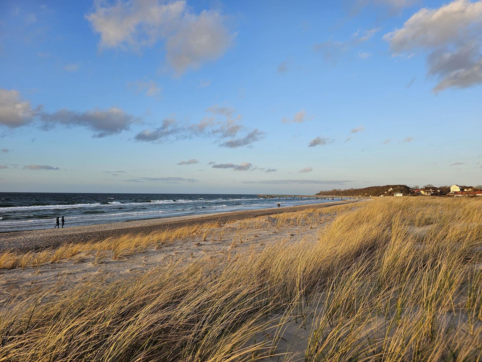 Strand mit Dünen und See, zwei Personen spazieren am Ufer entlang.