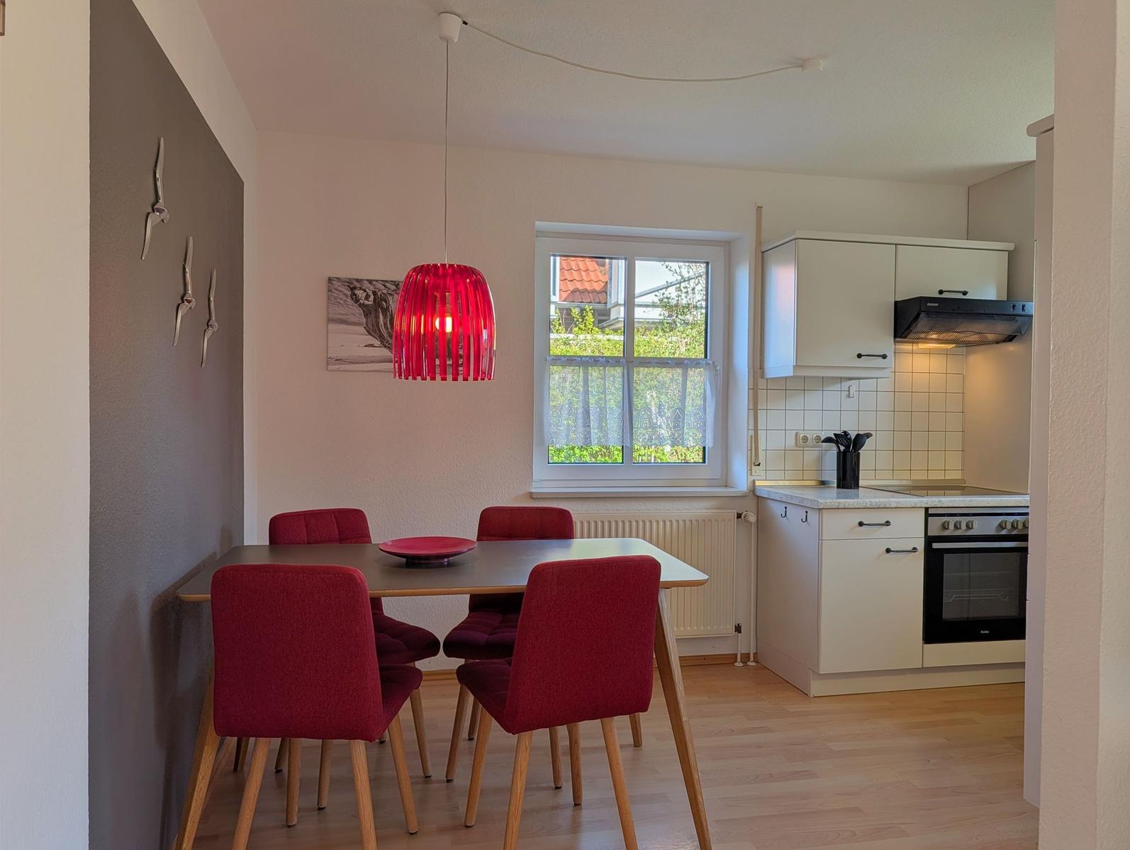 Essbereich mit roten Stühlen und Tisch. Küche mit weißen Schränken und Fenster.