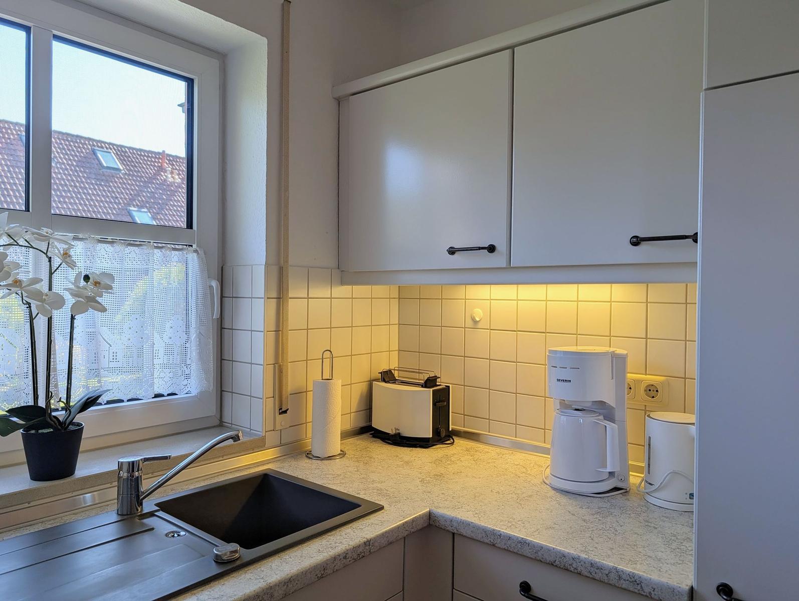 Küche mit Arbeitsfläche, Spüle, Kaffeemaschine und Toaster. Fenster mit weißen Vorhängen.