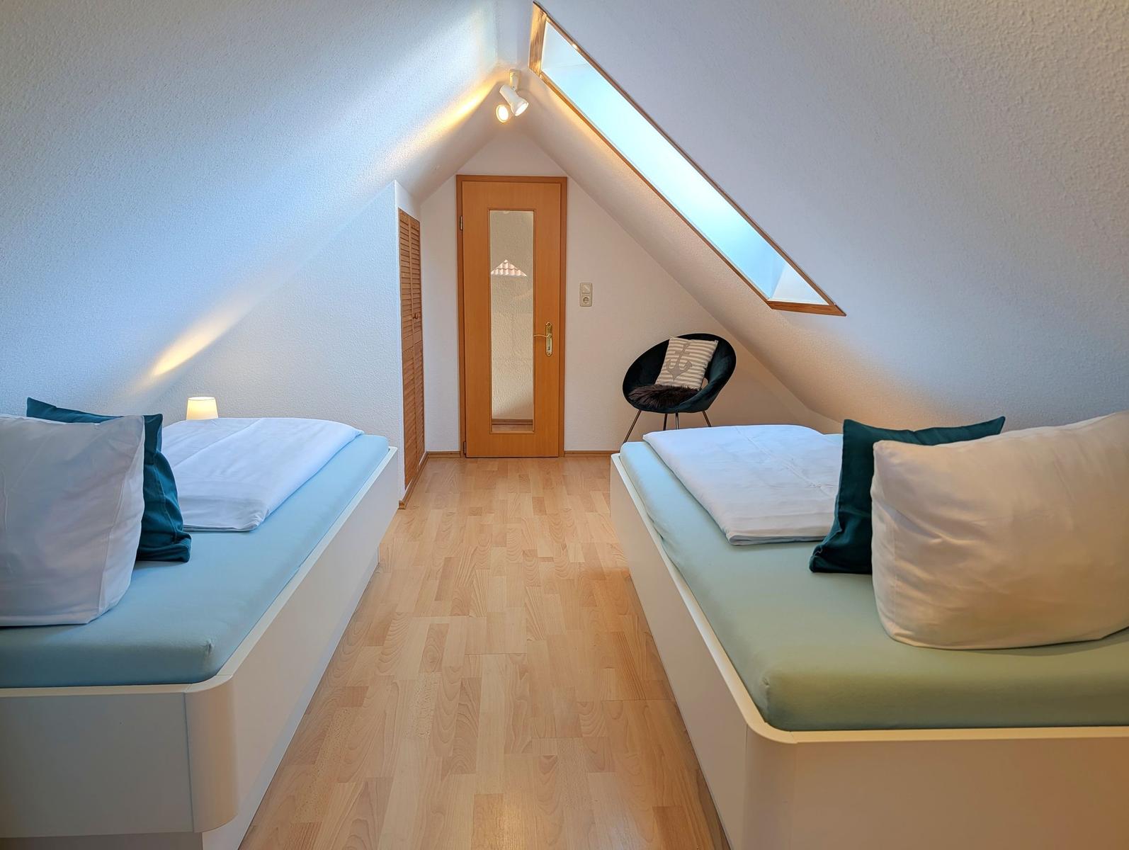 Two beds with pillows, wooden floor, skylight, and chair in attic room.