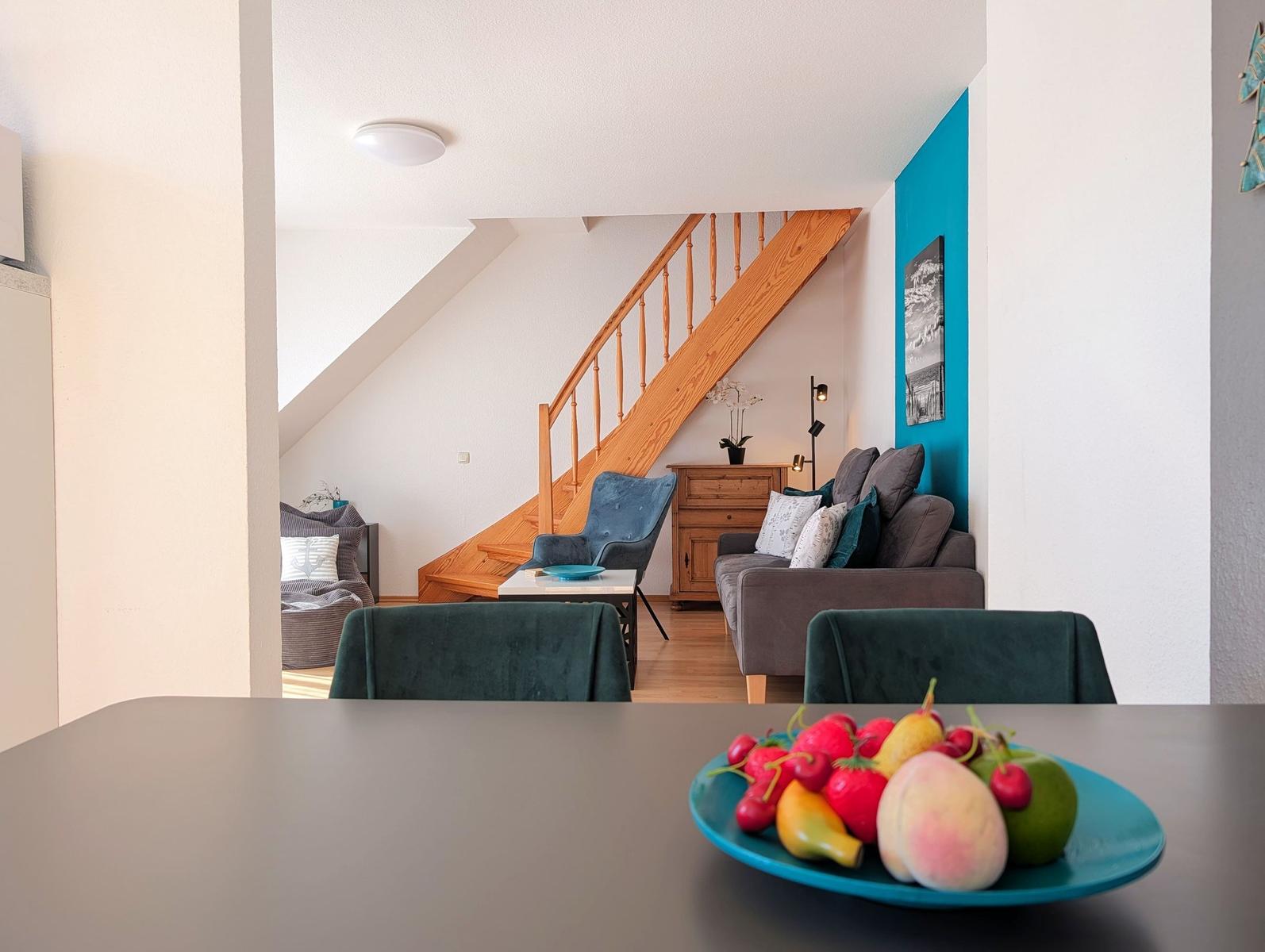 Dining area with fruit bowl, view into living room with wooden staircase and sofa.