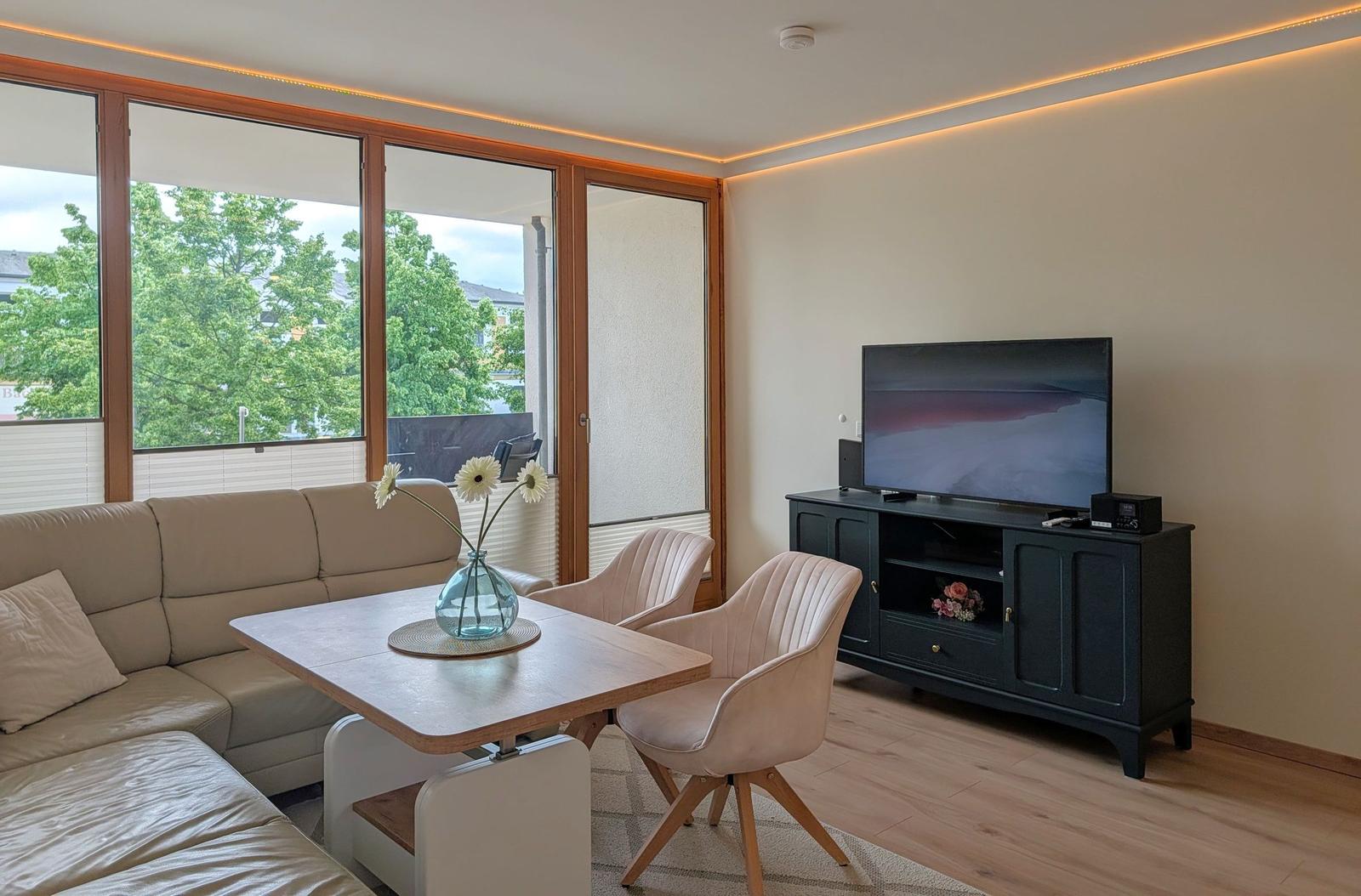 Wohnzimmer mit Sofa, Esstisch, Fernseher und großem Fenster mit Blick auf Bäume.