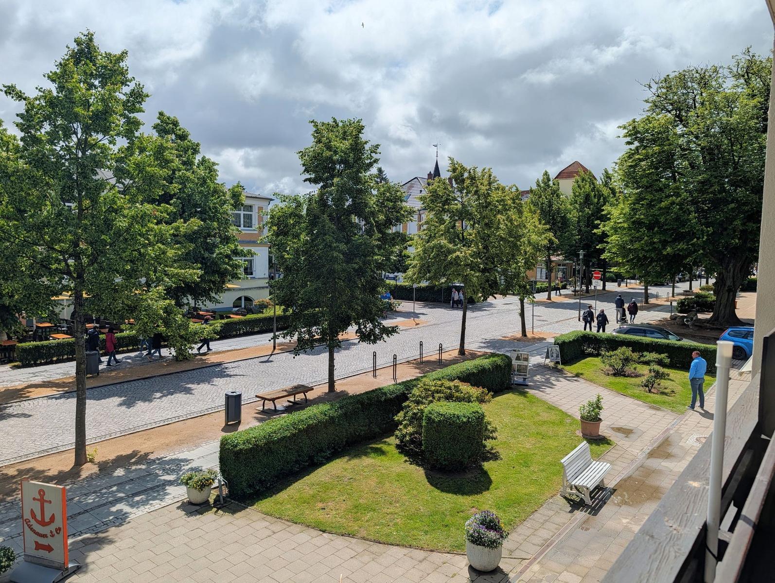 Aussicht auf grünen Platz mit Bäumen, Beeten und Gehweg. Menschen gehen und Autos parken.