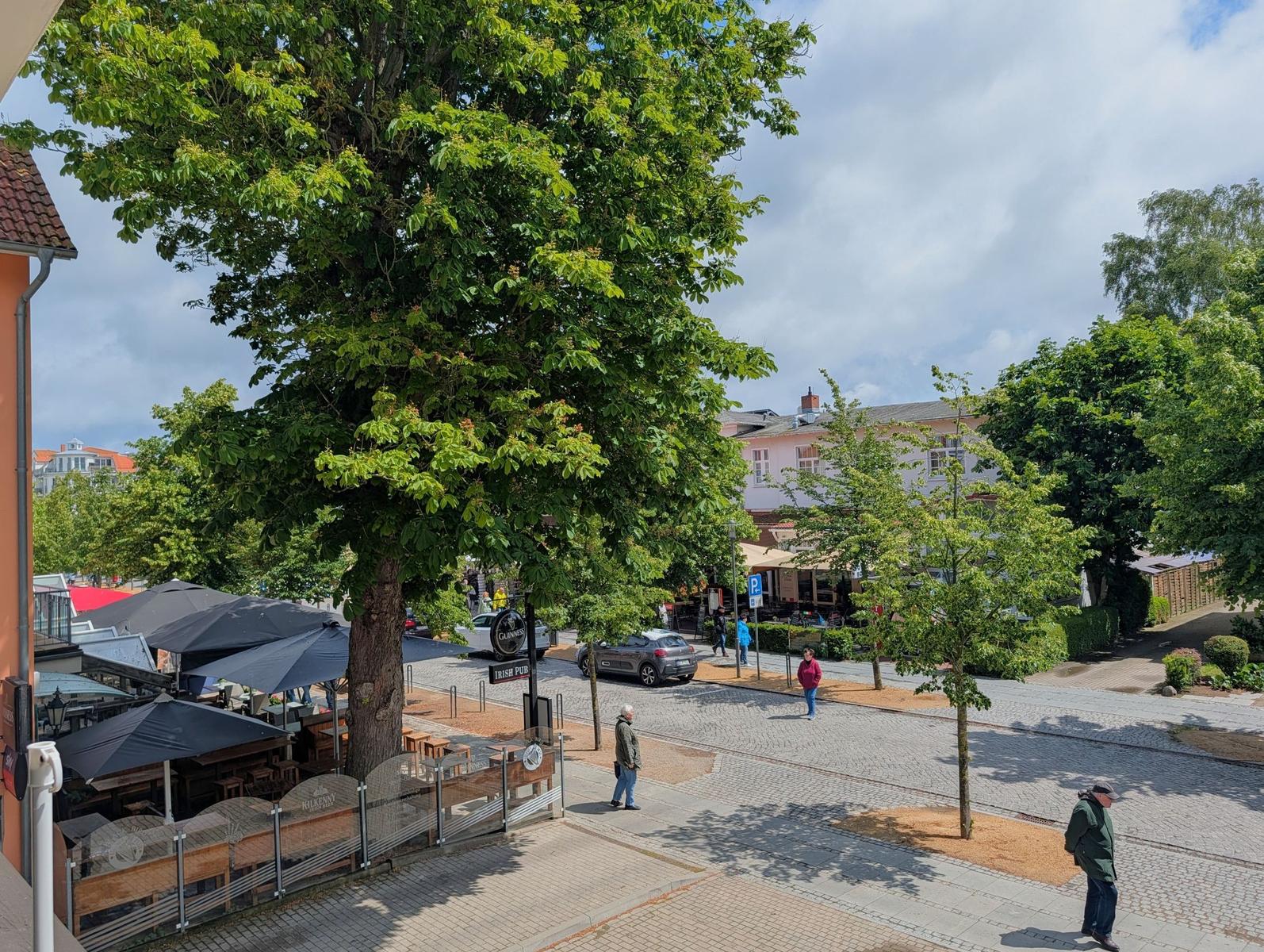 Straßenaussicht mit Bäumen, Café und Passanten. Steinpflaster und Parkplatz sind sichtbar.