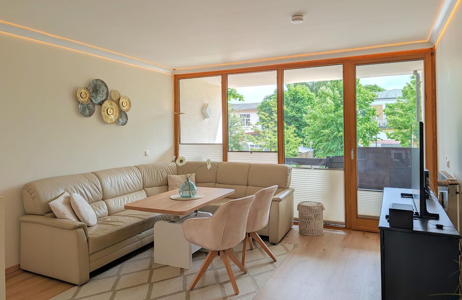 Wohnzimmer mit Sofa, Esstisch und Blick auf Balkon mit Bäumen.