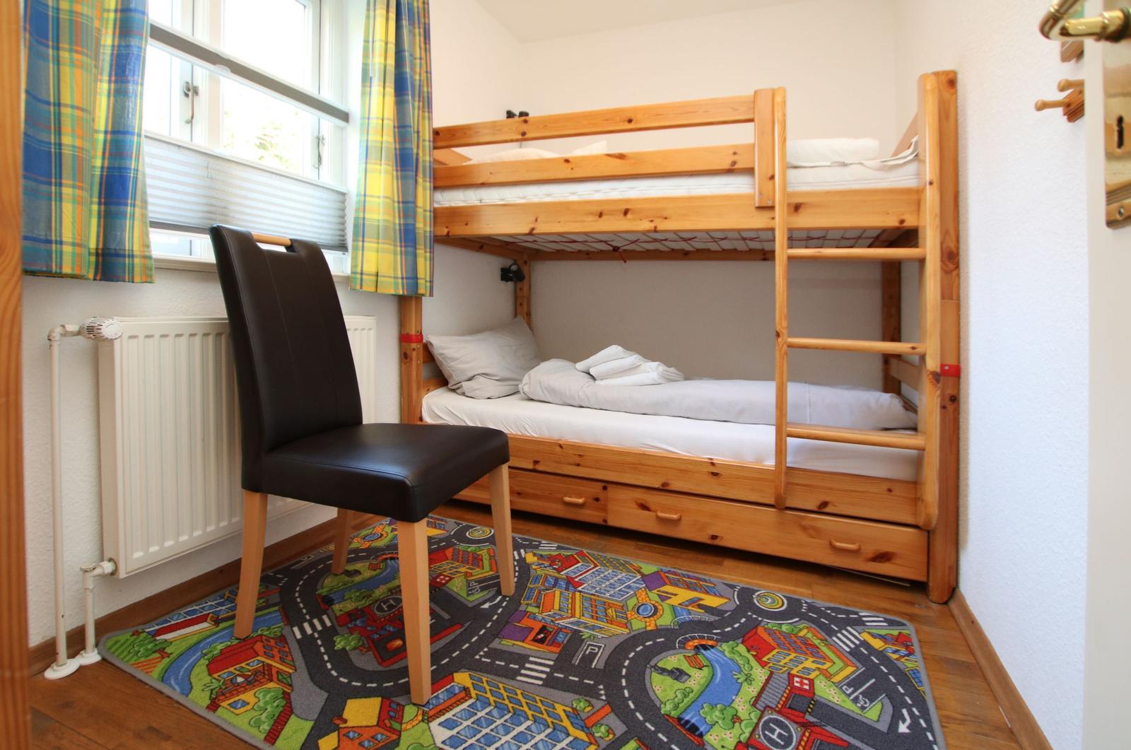 Ein Schlafzimmer mit Holz-Etagenbett, schwarzer Stuhl und buntem Kinder-Teppich.