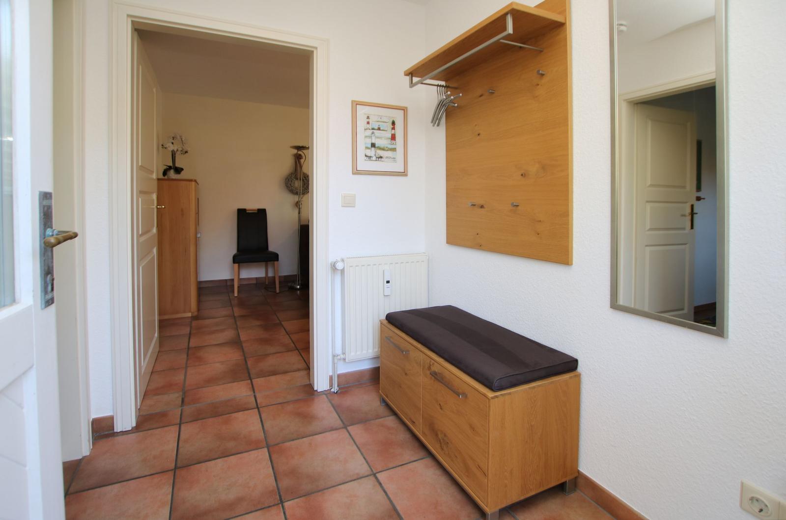 Eingangsbereich mit Holzbank, Wandgarderobe und Spiegel. Fliesenboden und weiße Wände.