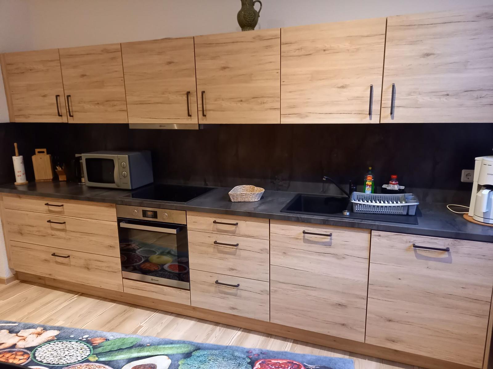 Küche mit Holzschränken, Spüle und Herd. Auf Arbeitsplatte stehen Geschirr und Küchenutensilien.