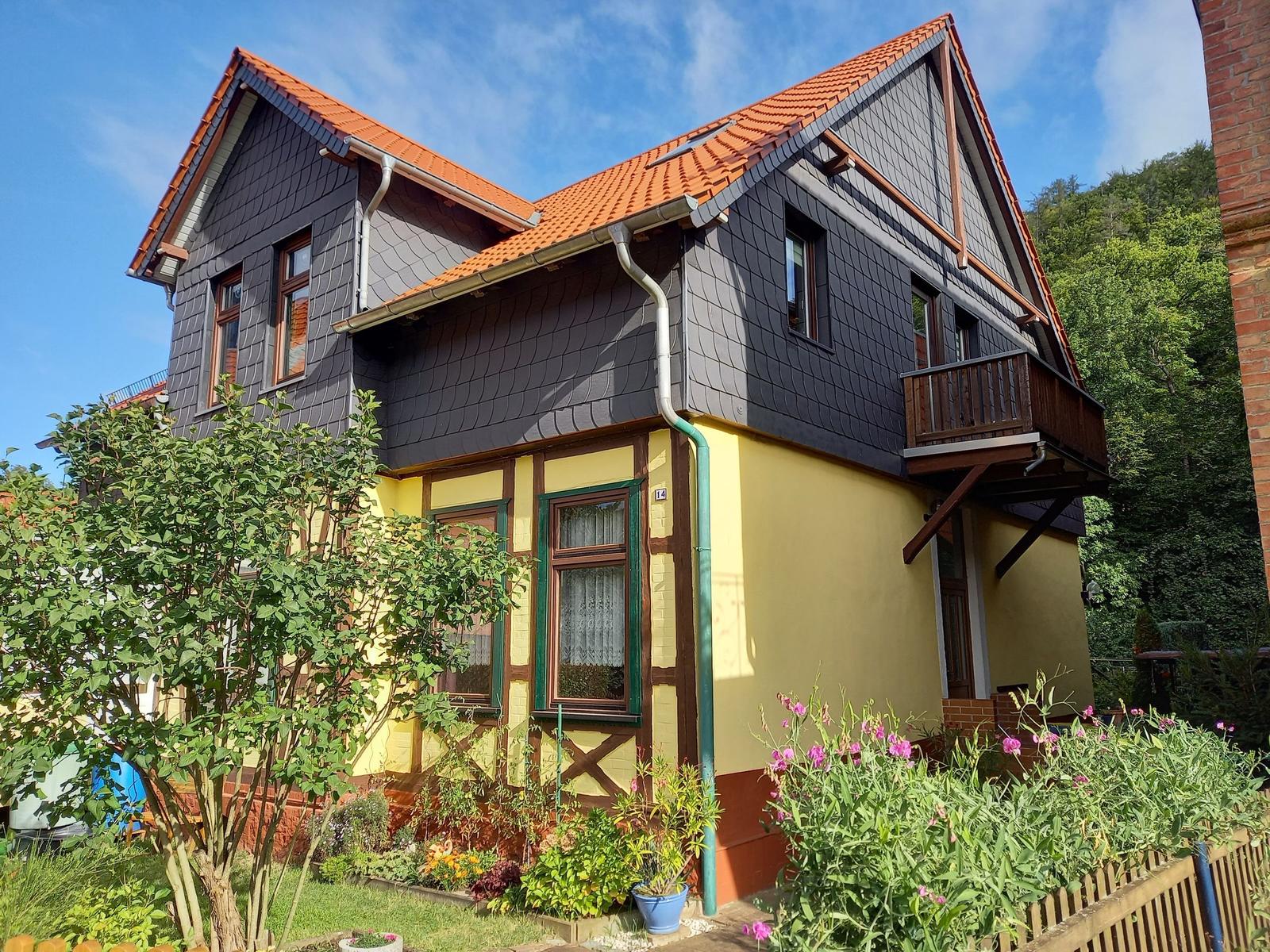 Haus mit gelbem Sockel und dunklem Dach, umgeben von Grün und Blumen.