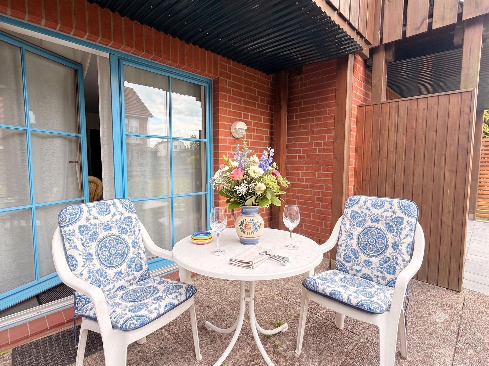 Terrasse mit Tisch, zwei Stühlen und Blumenvase. Hintergrund: rote Ziegelwand und blaue Fenster.