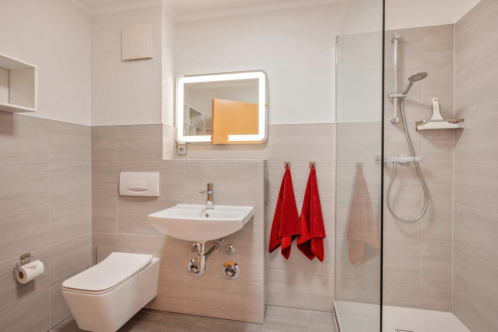 Badezimmer mit Dusche, Waschbecken und Toilette. Rote Handtücher hängen an der Wand.
