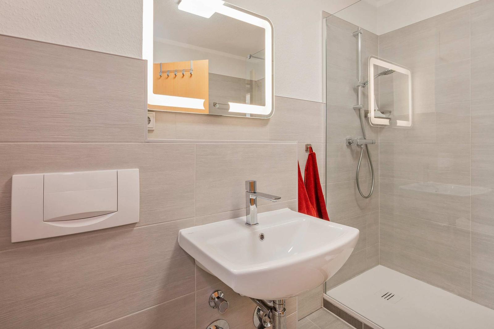 Badezimmer mit Dusche, Waschbecken und beleuchtetem Spiegel