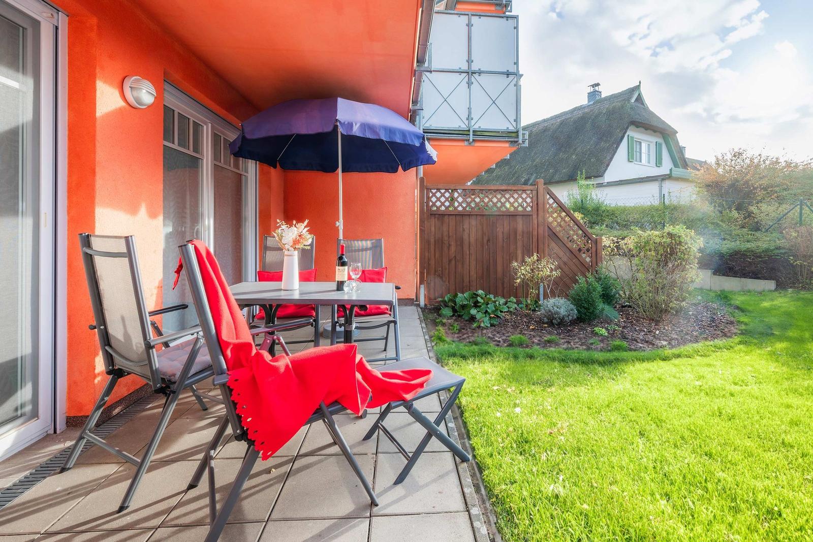 Terrasse mit Tisch, Stühlen und Sonnenschirm. Grünfläche und Zaun im Hintergrund.