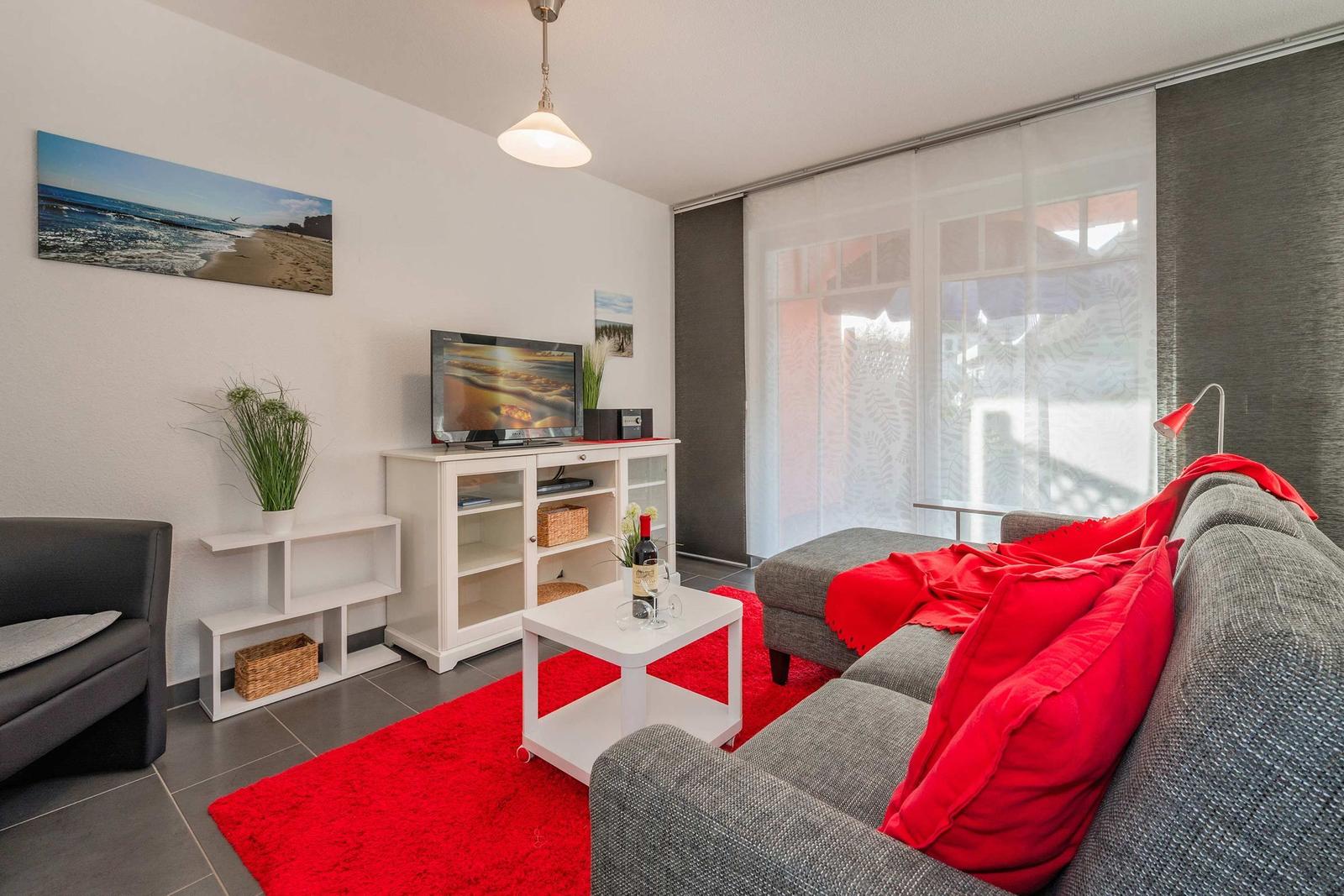 Wohnzimmer mit grauem Sofa, rotem Teppich und Fernseher auf weißen Möbeln.