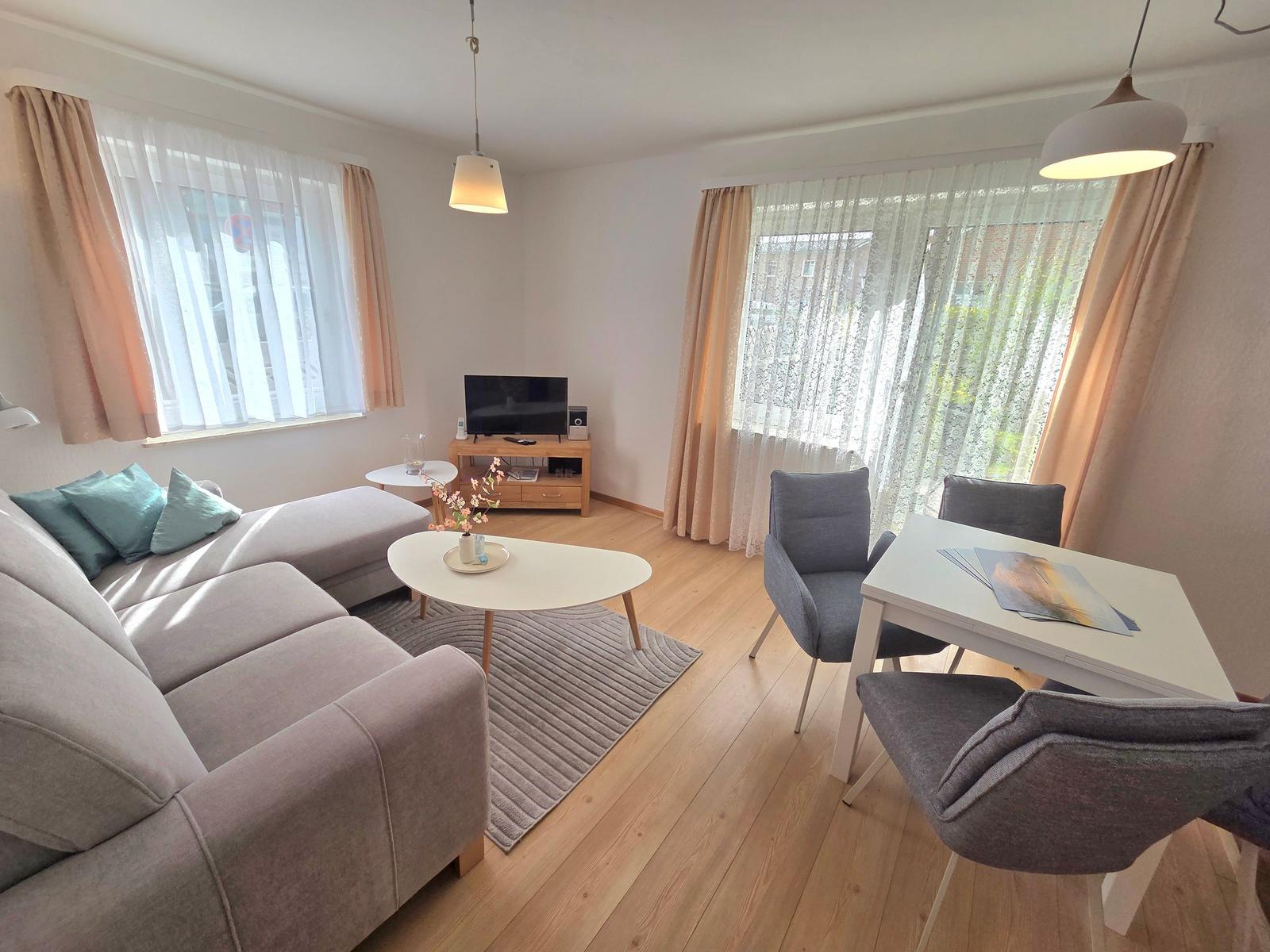 Wohn- und Essbereich mit Sofa, Tisch und Stühlen. Holzboden und Fenster mit Vorhängen.