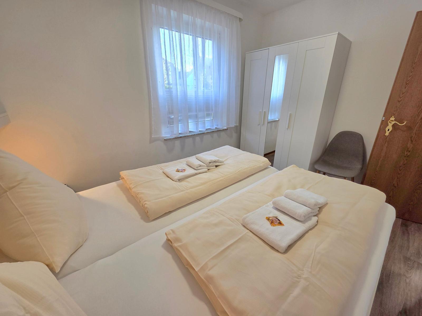 Zimmer mit Doppelbett, Kleiderschrank und Fenster mit Vorhängen.