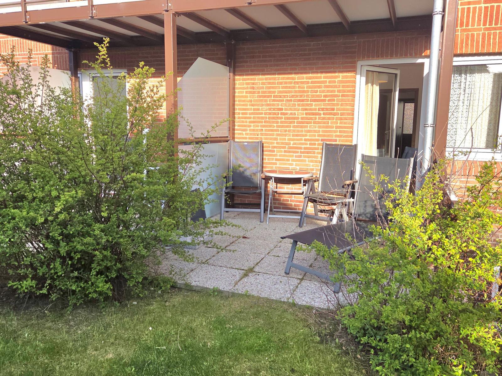 Terrasse mit Sitzgelegenheiten und Garten. Hintergrund: Backsteinmauer und offene Tür.