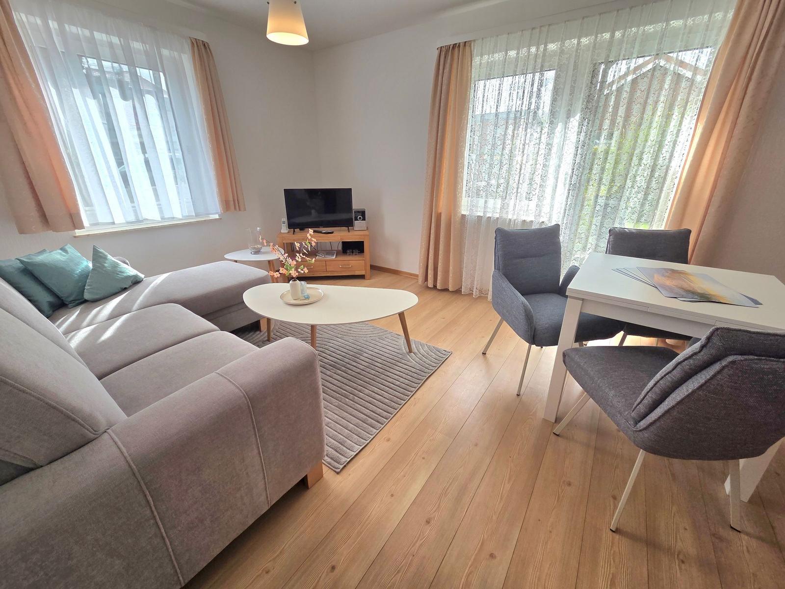 Wohnzimmer mit Sofa, Esstisch und Fernseher. Holzboden und große Fenster.