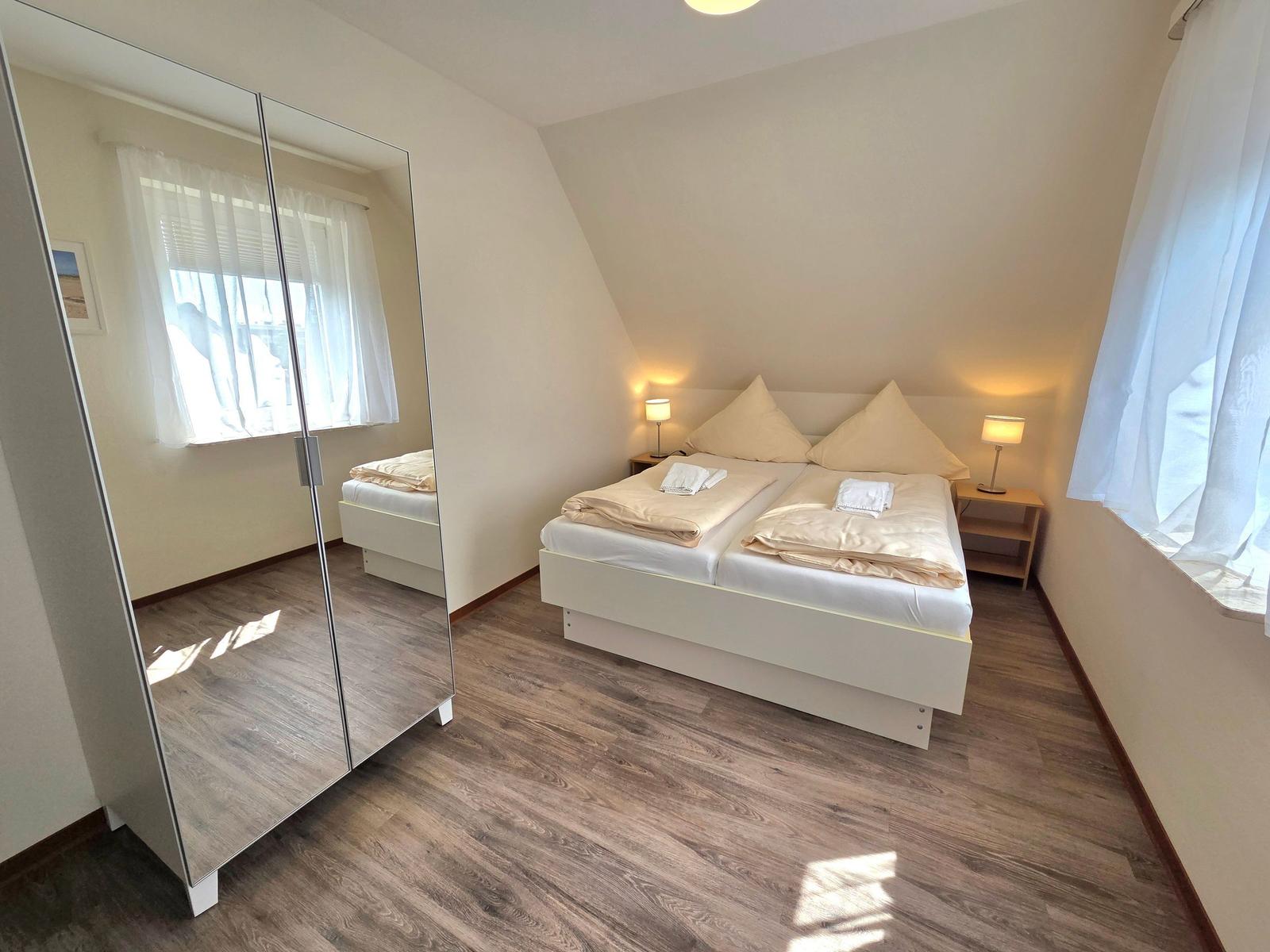 Schlafzimmer mit Doppelbett, zwei Nachttischen und Spiegelschrank. Holzdielenboden und Fenster mit Vorhängen.