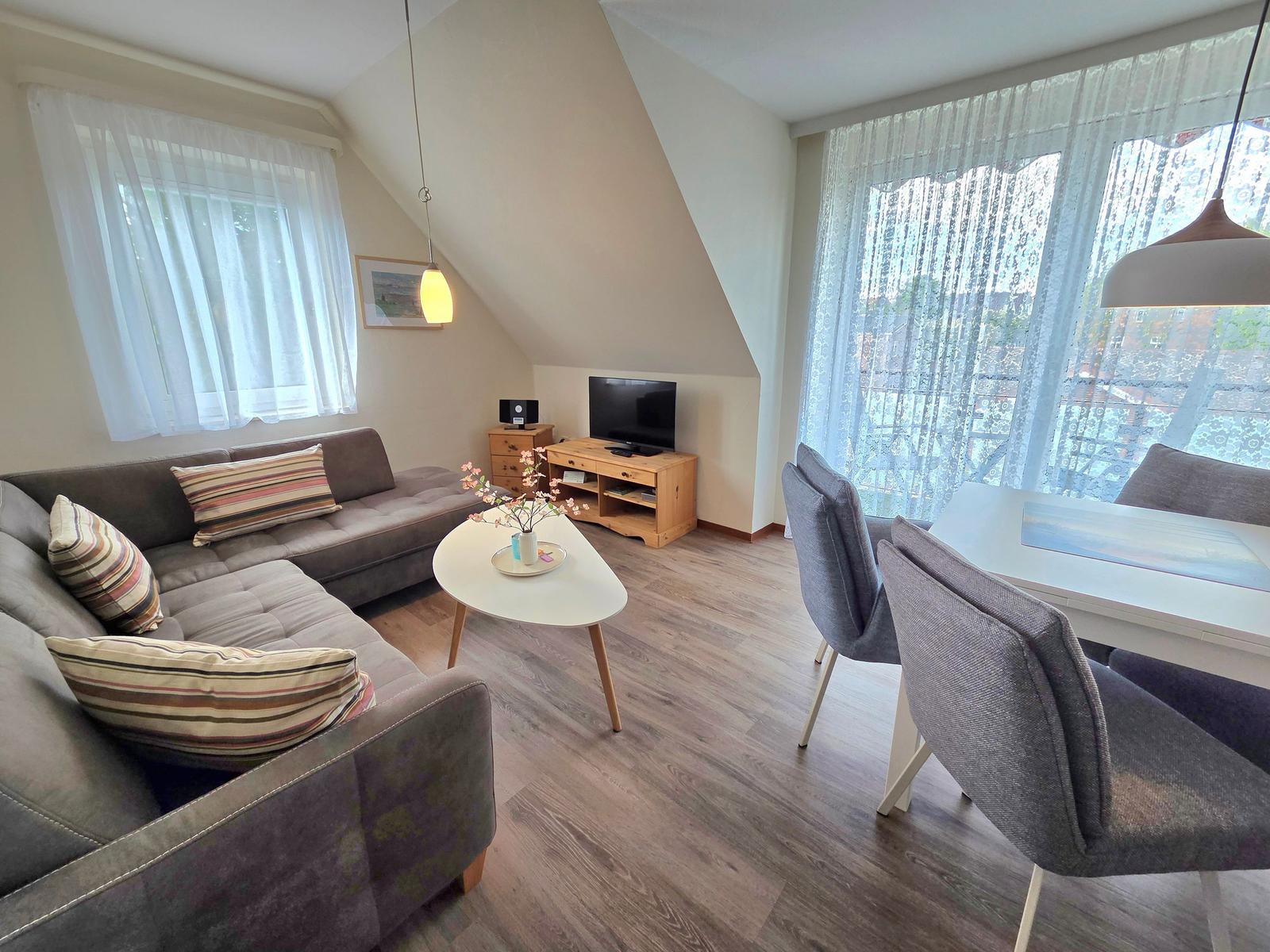 Wohn- und Essbereich mit Sofa, Tisch und TV. Holzboden und große Fenster.
