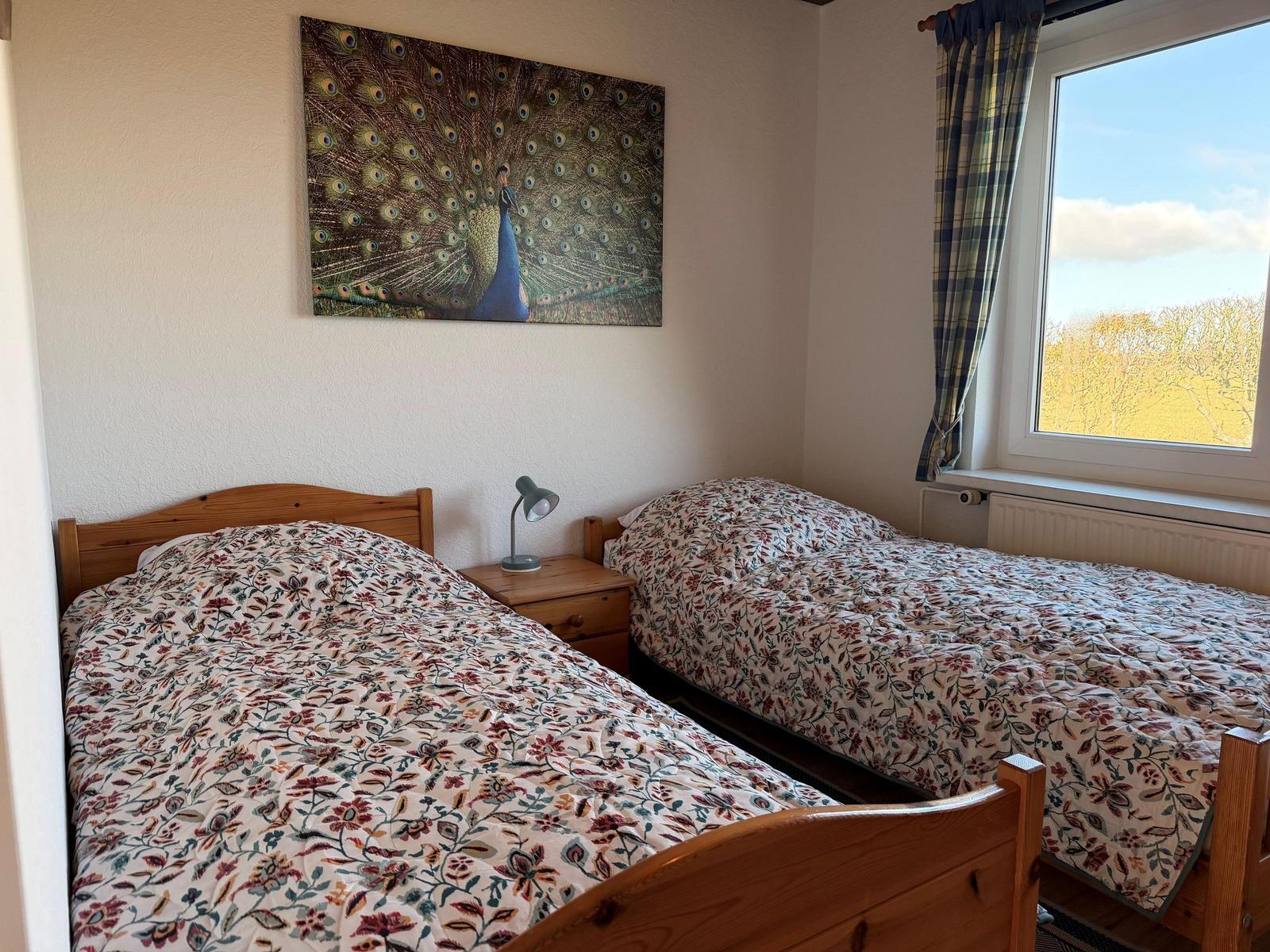 Zwei Einzelbetten mit floraler Bettwäsche, Wandbild eines Pfauens und Fenster mit Blick.