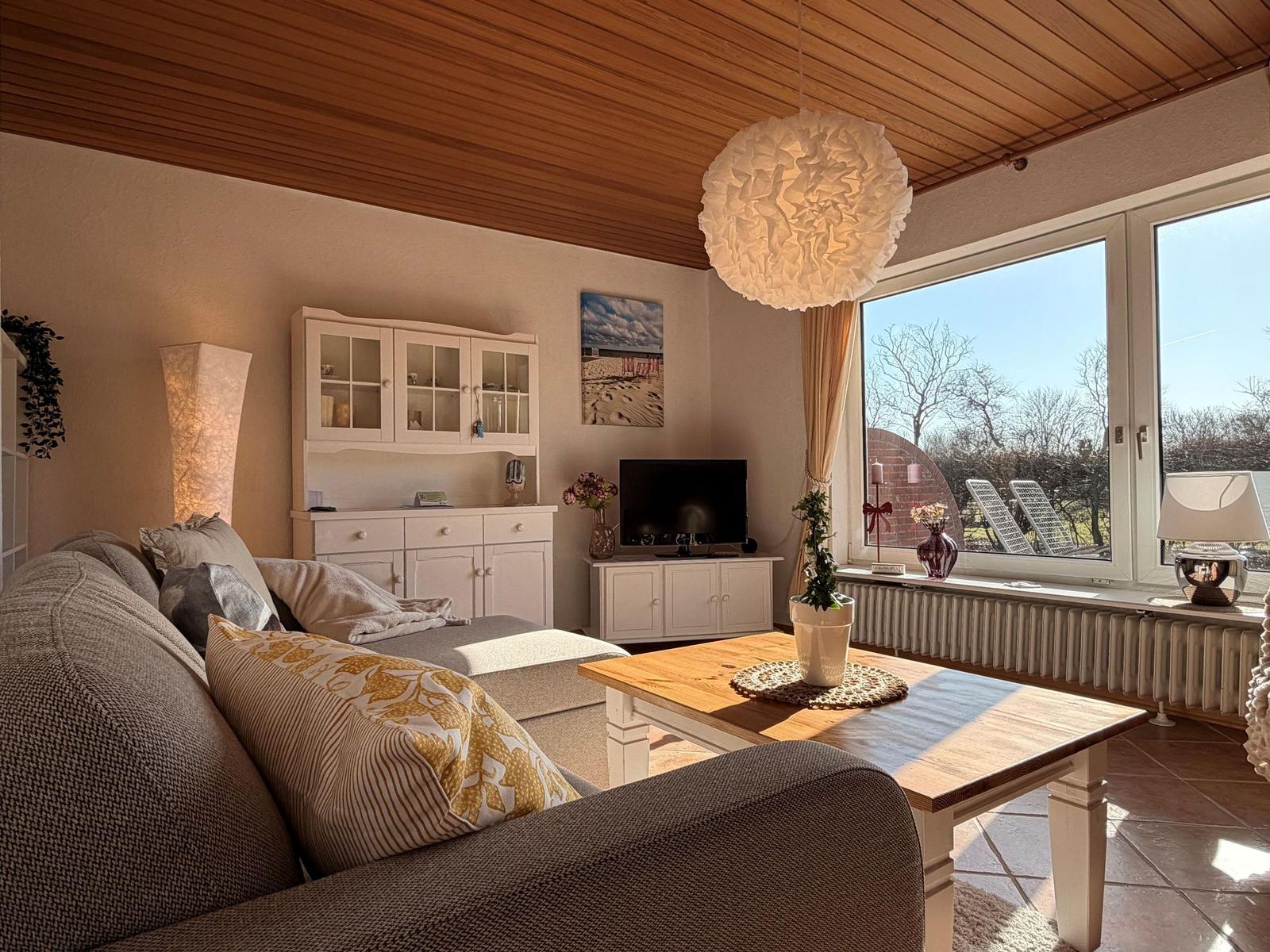 Wohnzimmer mit Sofa, Couchtisch und TV. Große Fenster mit Blick auf Garten.