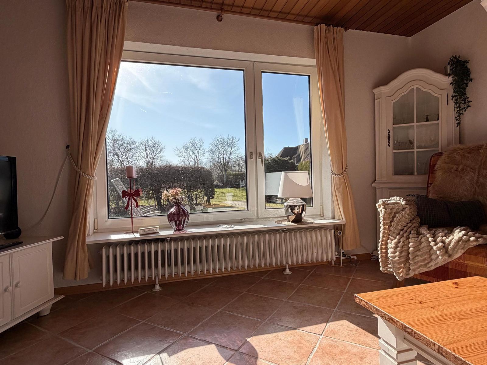 Wohnzimmer mit großem Fenster, Holzdecke, Schrank und Sessel. Blick auf Garten und Himmel.