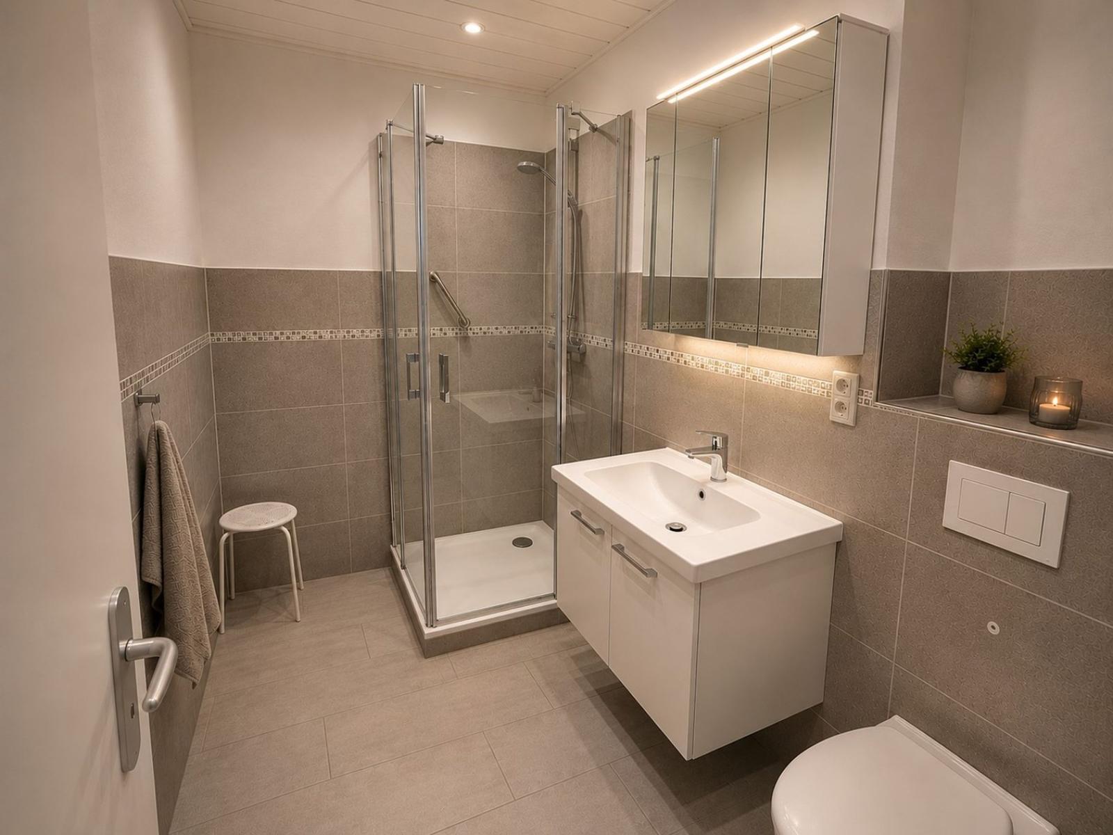 Badezimmer mit Dusche, Waschbecken und Toilette. Fliesenwand, Spiegel und kleine Pflanze.