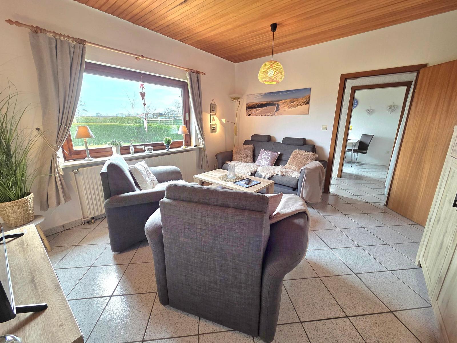 Wohnzimmer mit Sofa, Sesseln und großem Fenster. Holzdecke und Fliesenboden.