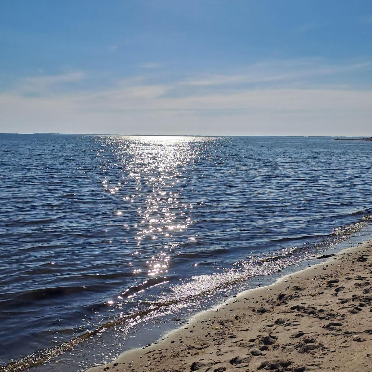Strand mit sanften Wellen und Sonnenreflexionen auf dem Wasser unter blauem Himmel.