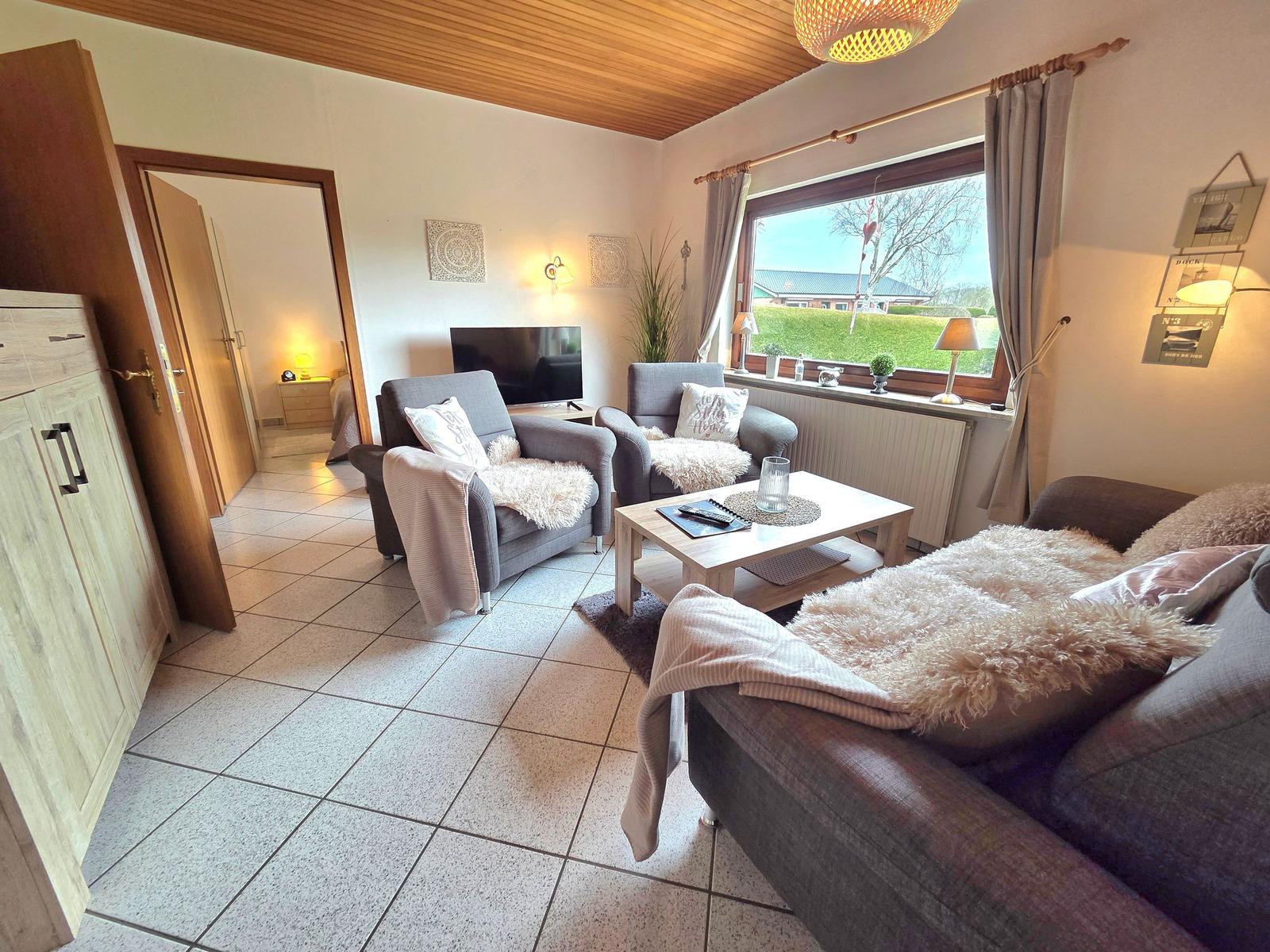 Wohnzimmer mit Sofa, Sesseln, Couchtisch und TV. Fenster mit Blick auf Grünfläche.